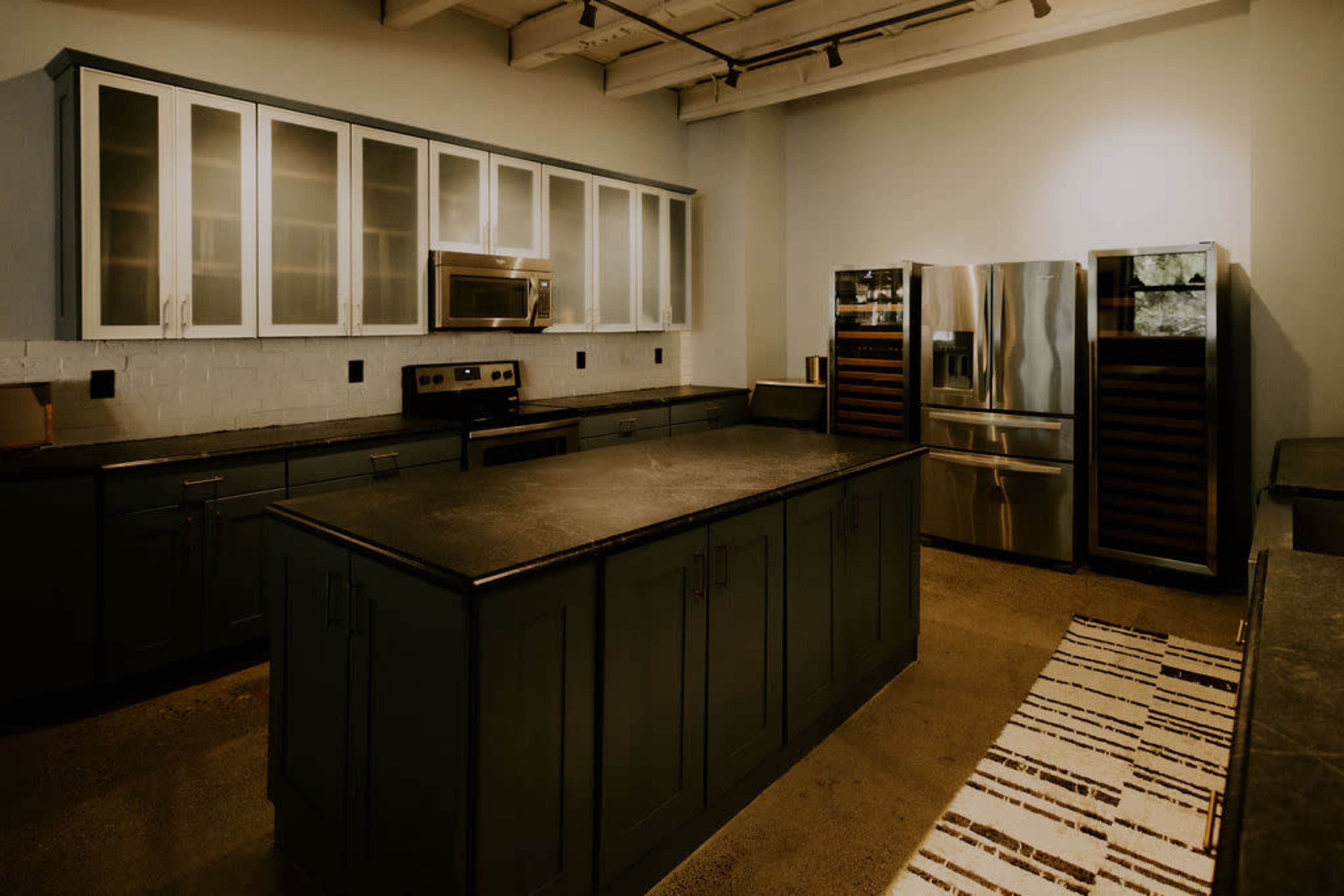 A modern kitchen with a central island, dark cabinetry, stainless steel appliances, and a patterned rug on the floor.