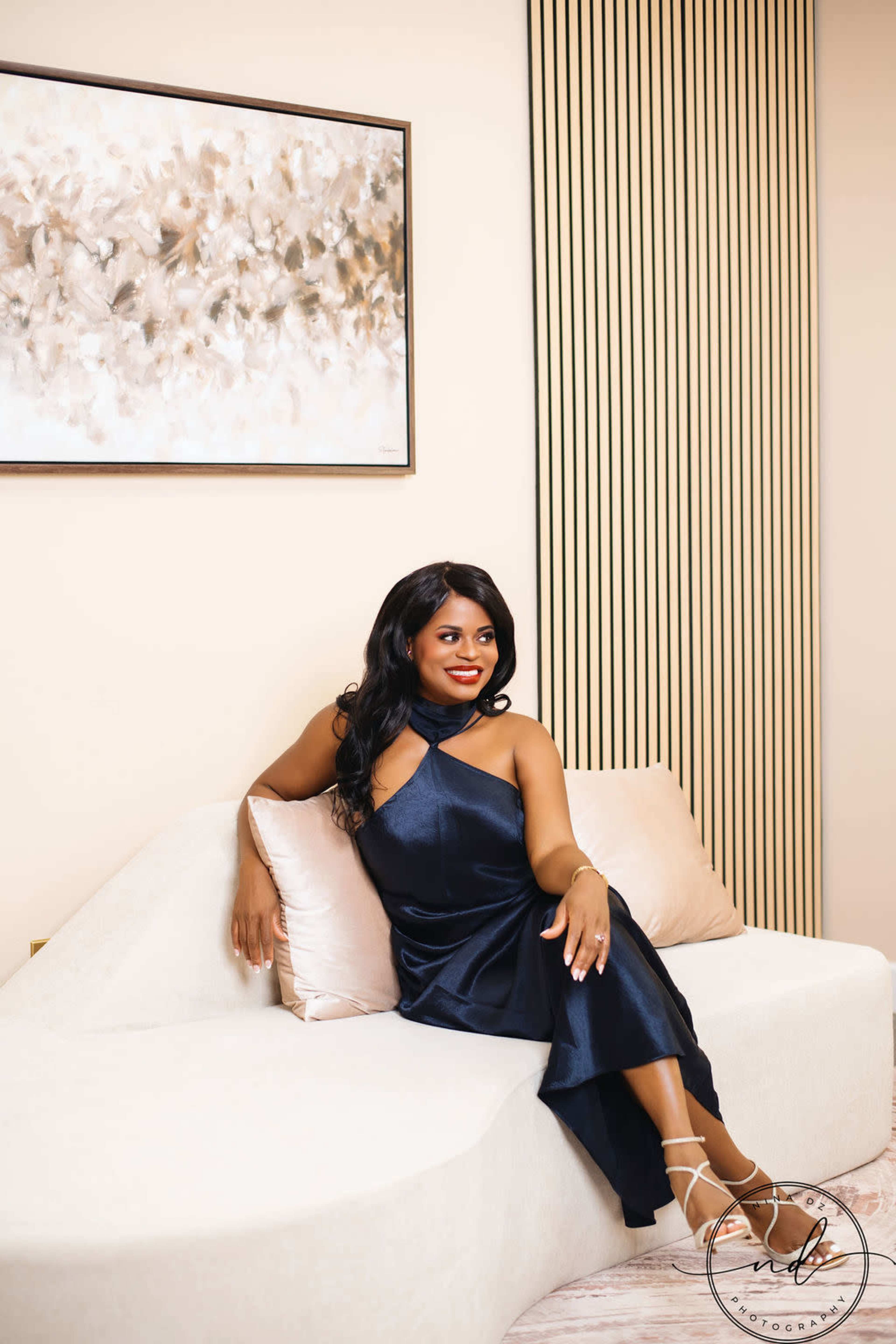 A woman in a navy blue dress sits elegantly on a curved sofa in a well-decorated interior space.