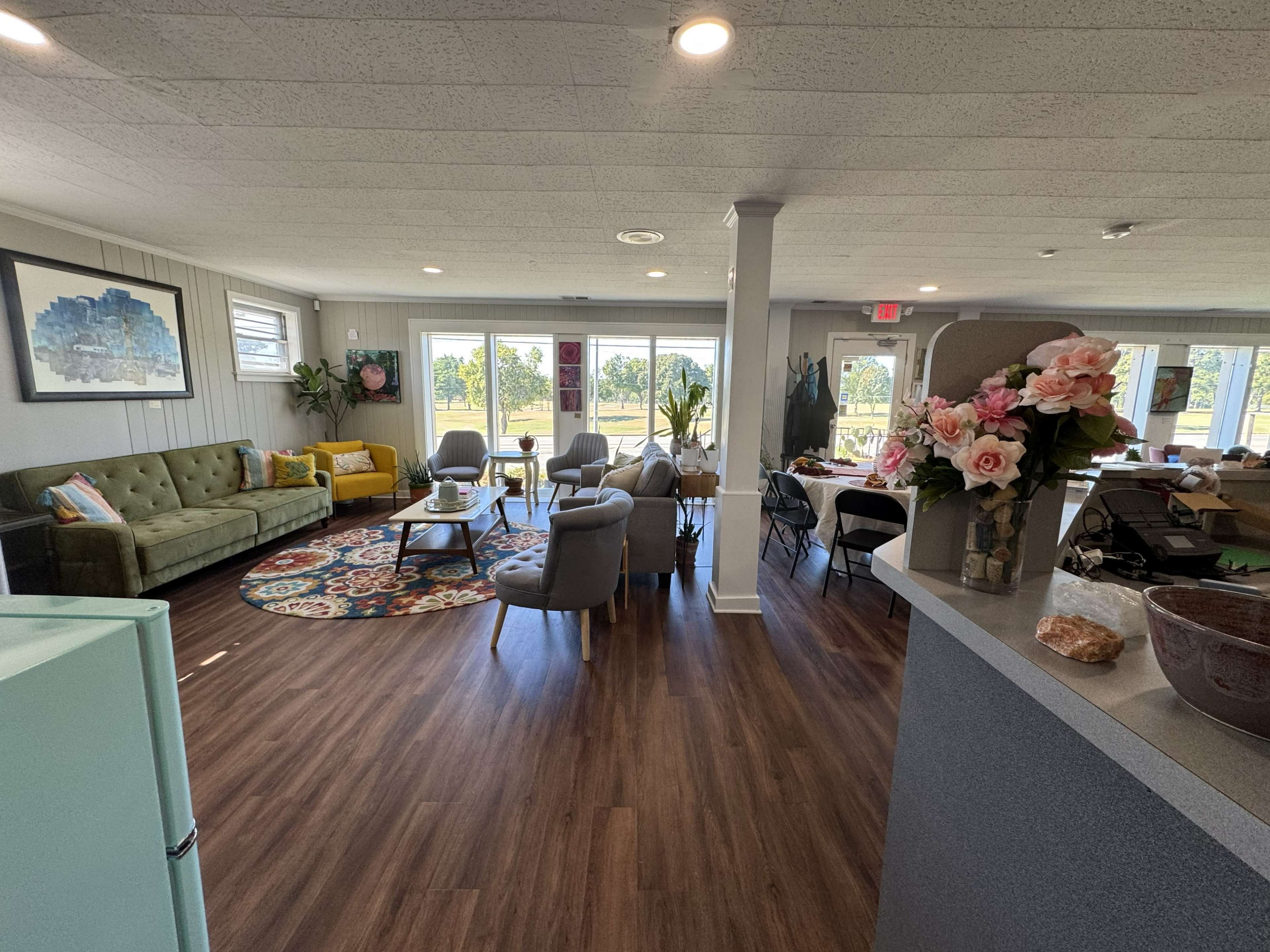 The image shows a spacious room with a mix of seating arrangements, large windows providing natural light, and decorative plants, along with a kitchenette area in the foreground.