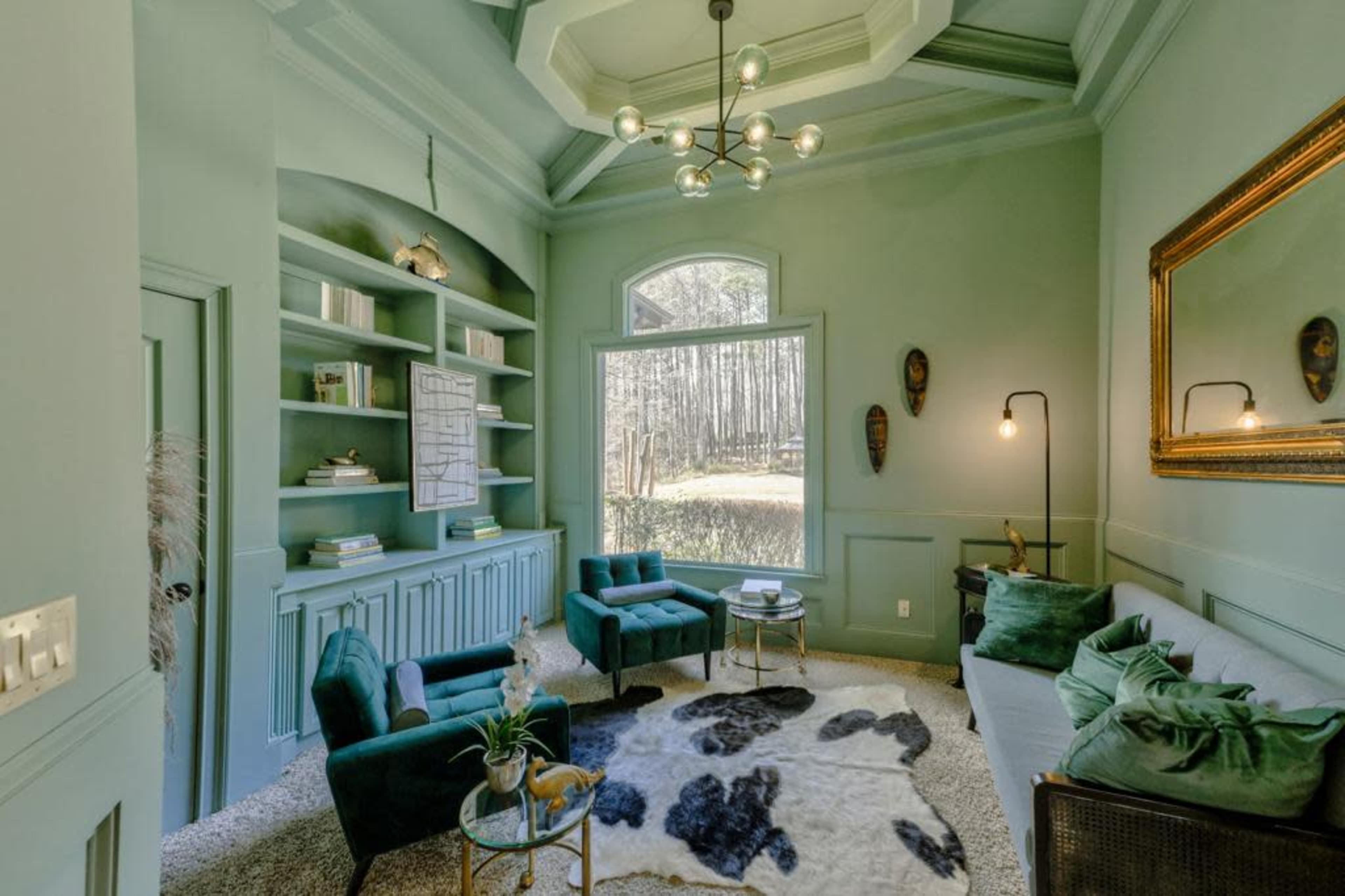 A cozy living space features teal walls, a plush carpet, a large window overlooking trees, and various seating arrangements.