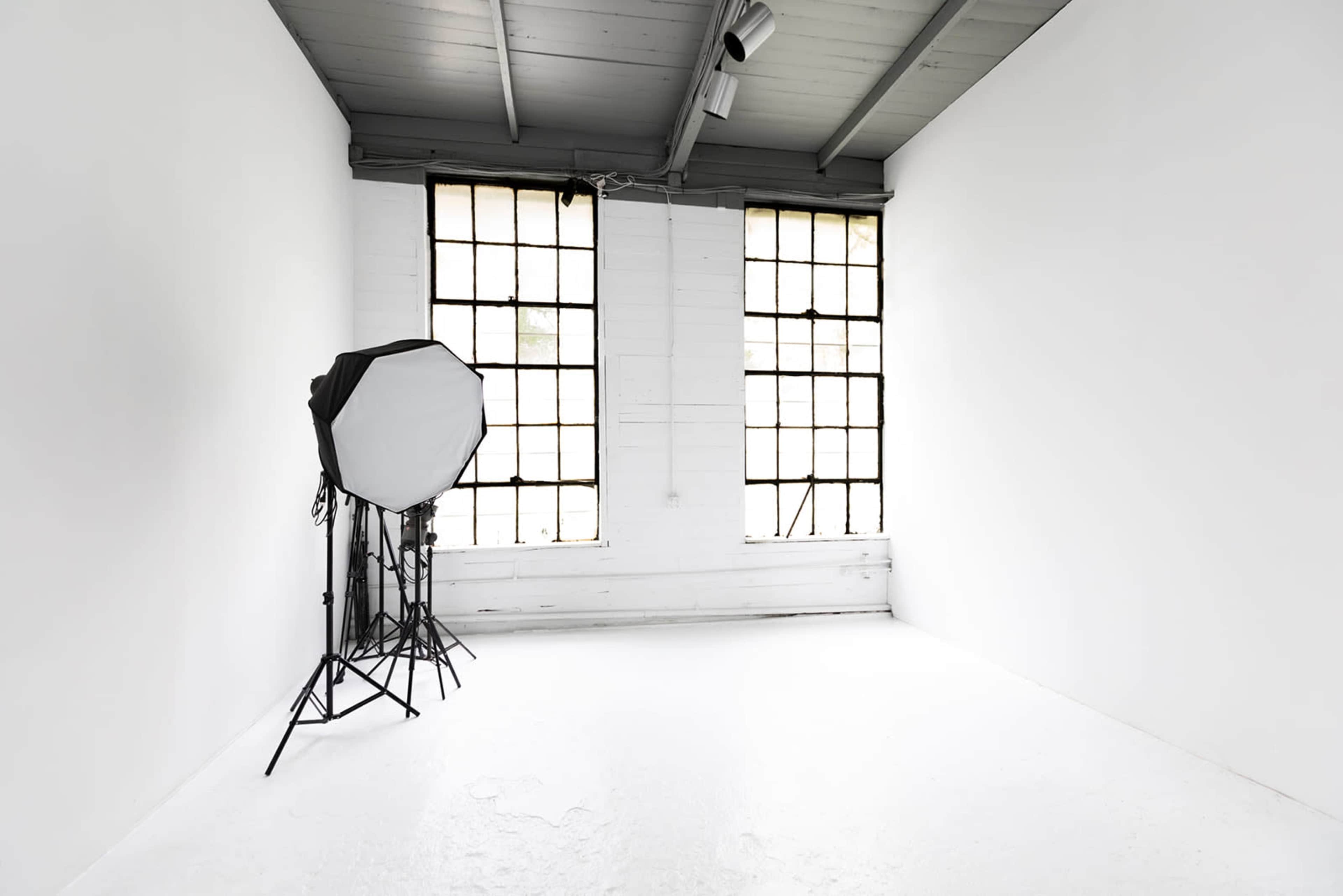 The image shows a minimalist white room with two large windows and a softbox lighting setup in one corner.