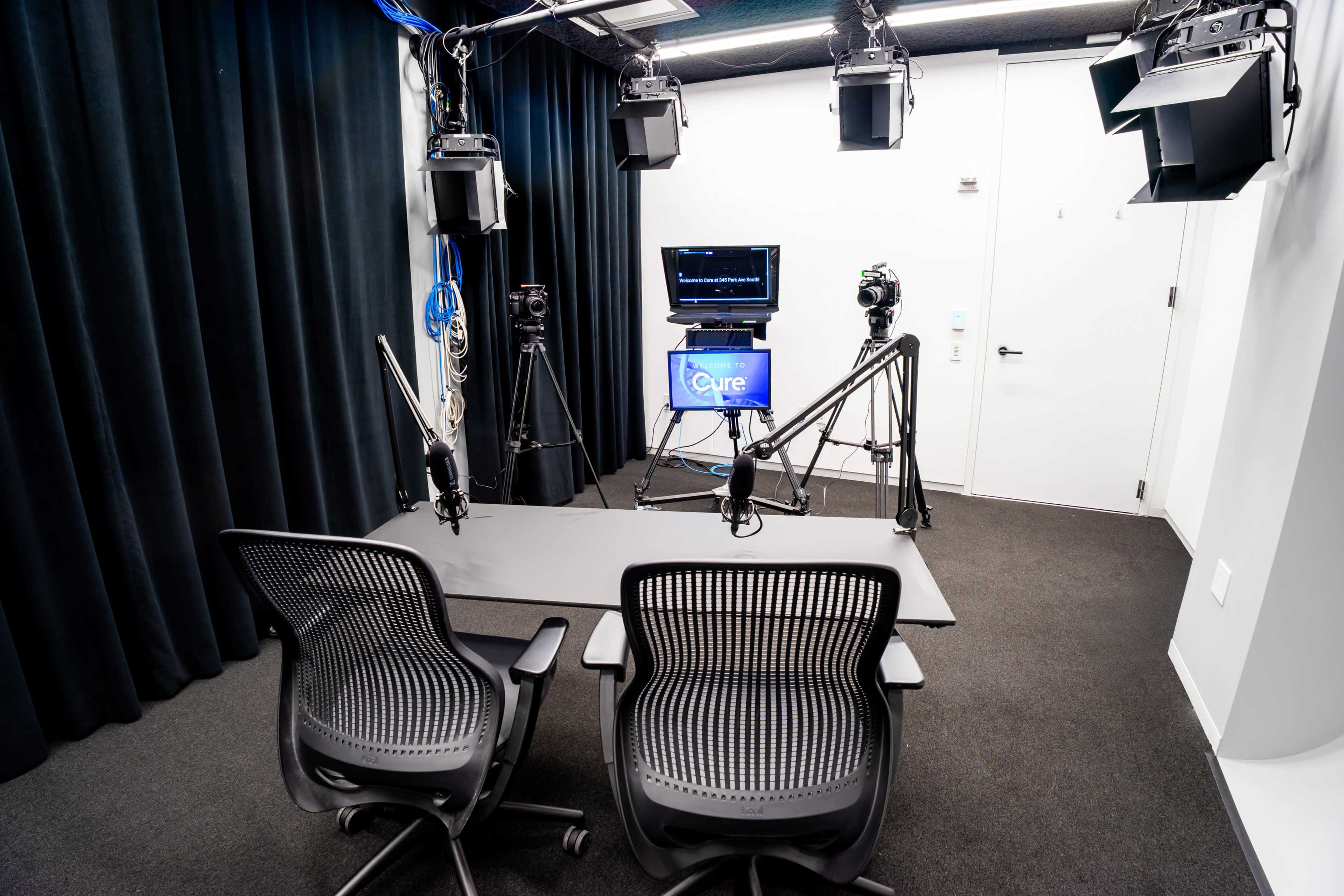 The image shows a video production studio equipped with two chairs, a large table, and multiple mounted lights and cameras.