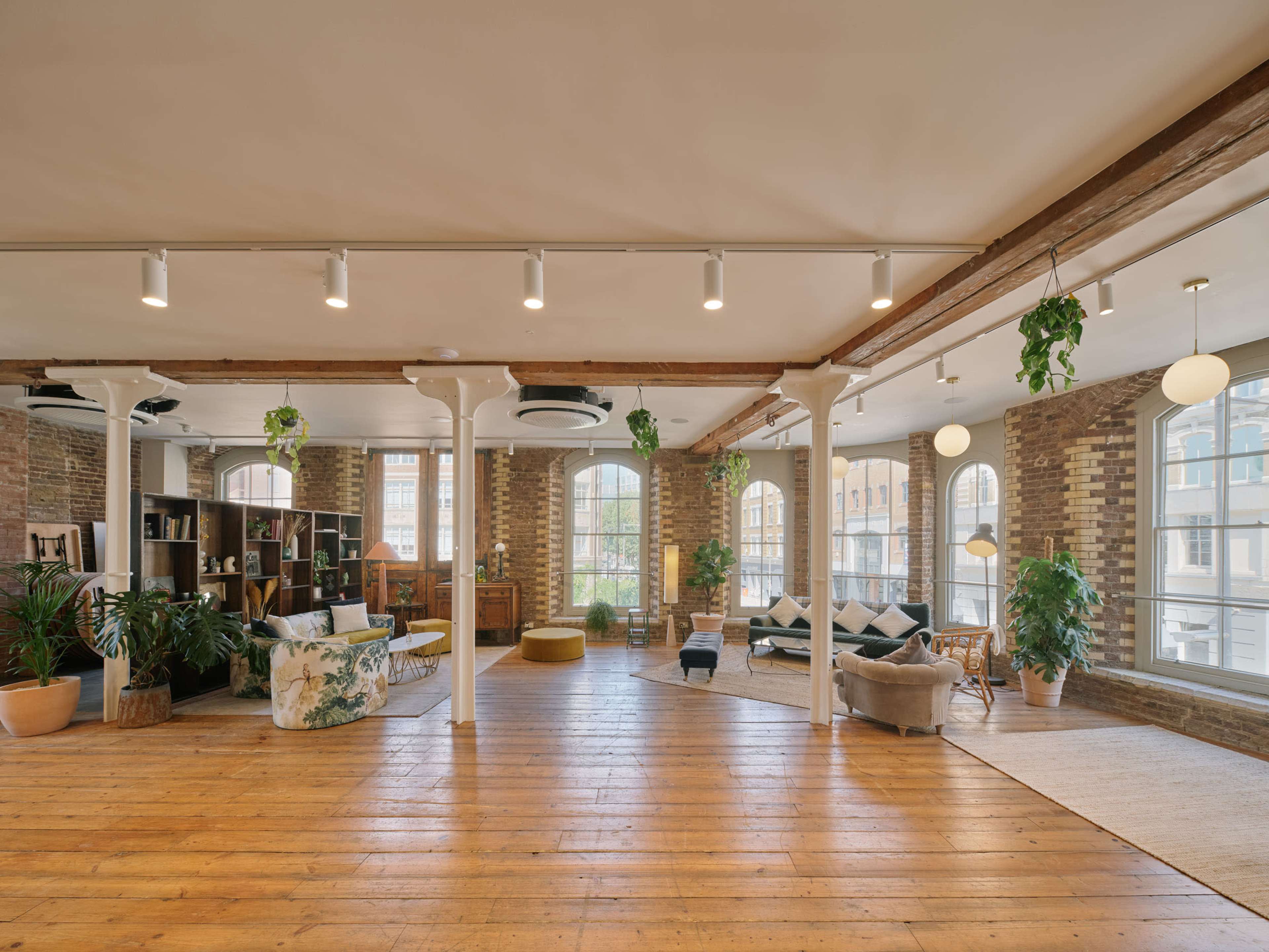 The image shows a spacious, well-lit loft with wooden flooring, exposed brick walls, large windows, and various plants, featuring a mix of seating areas and bookshelves.
