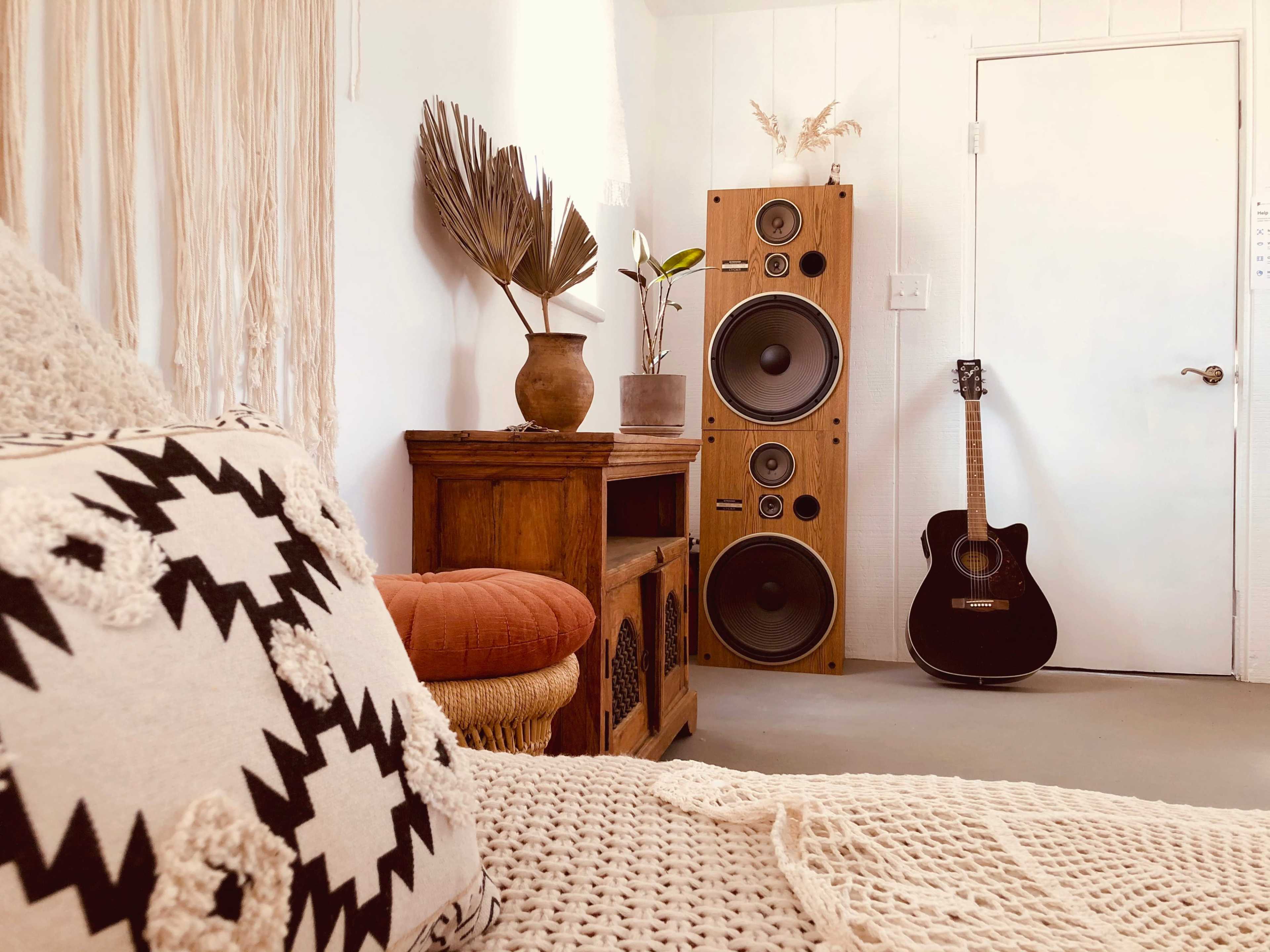 A cozy room features a bed with patterned pillows, a wooden cabinet, a large speaker setup, a guitar, and a potted plant.