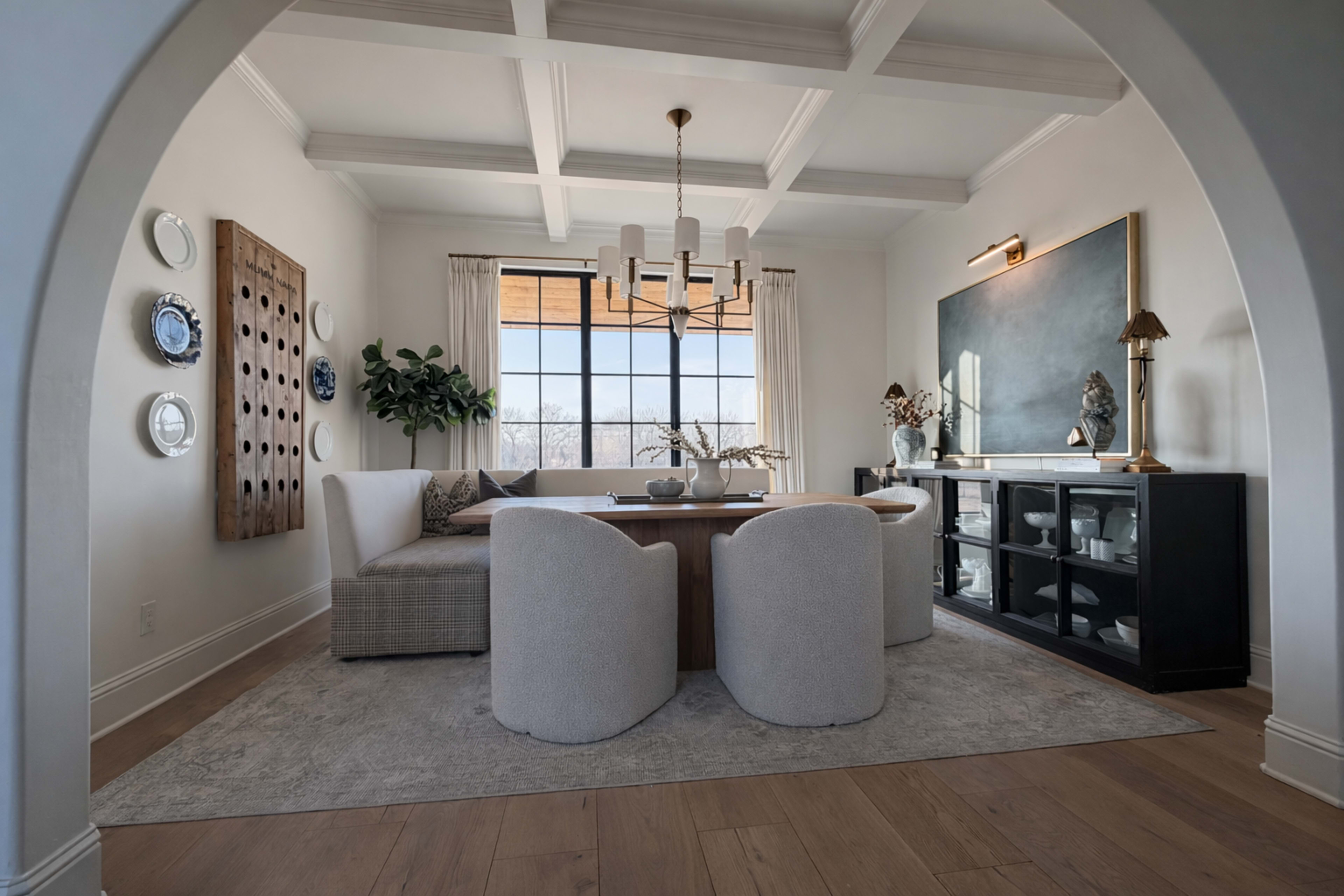 The image depicts a modern dining room featuring a large table surrounded by circular chairs, a sectional sofa, and large windows allowing natural light to illuminate the space.