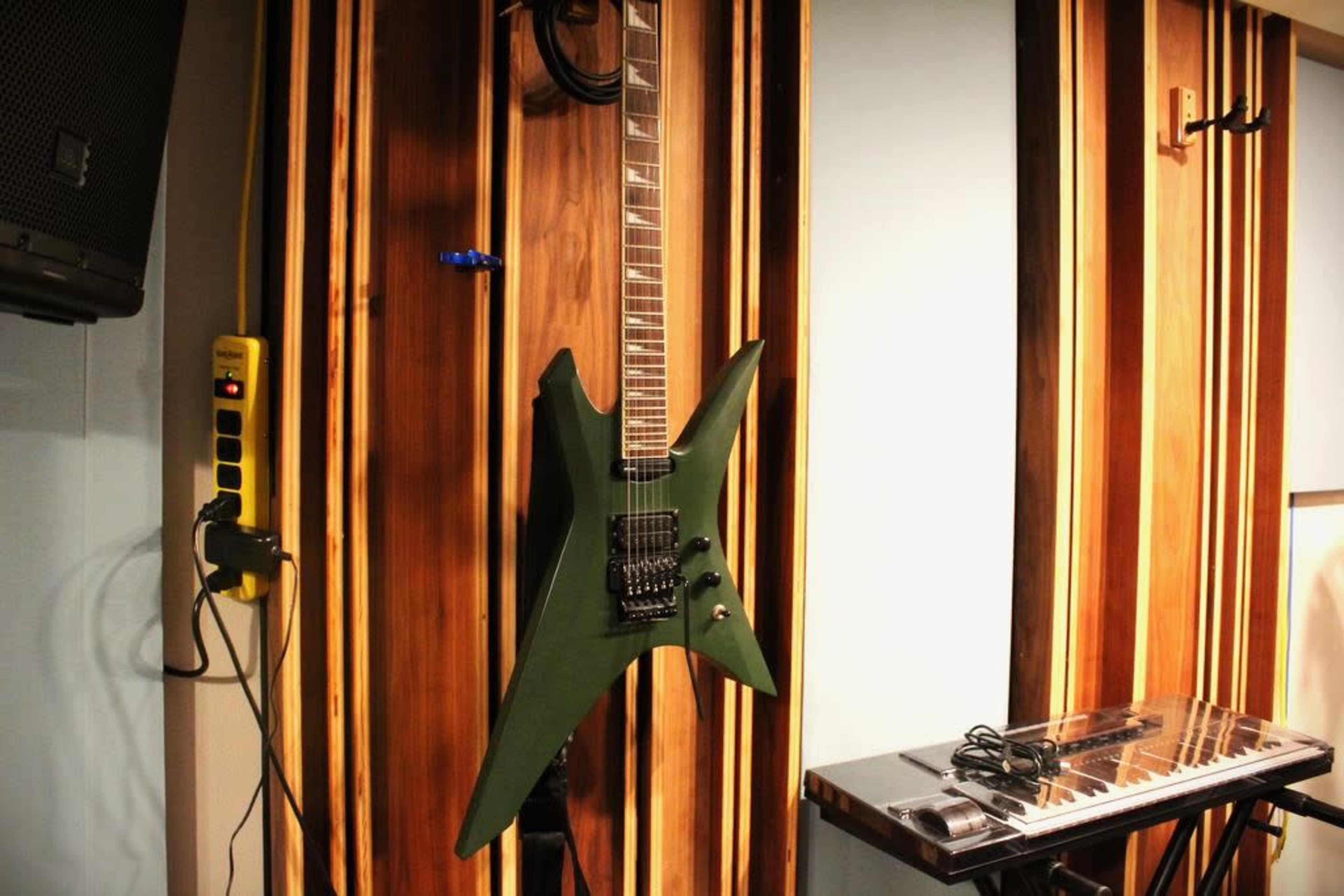 A green electric guitar with a distinctive shape is hanging on a wooden wall next to a keyboard and a power outlet.