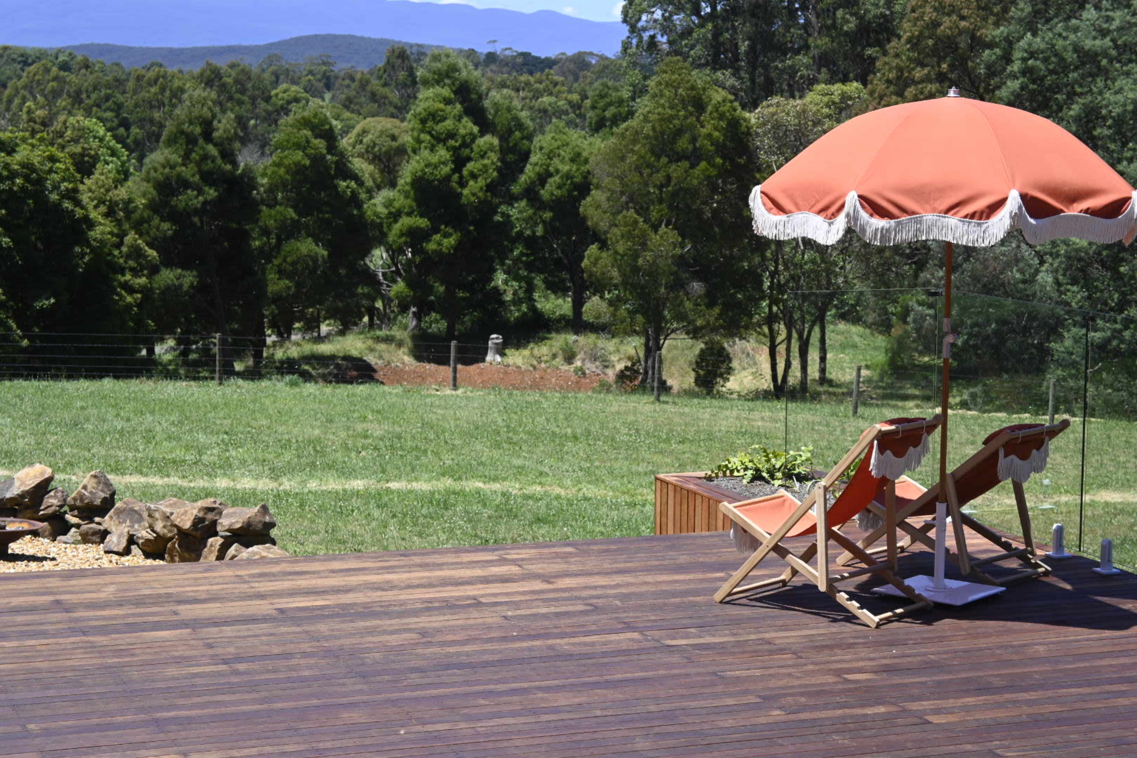 A wooden deck features two lounge chairs under a red umbrella, overlooking a grassy area with trees and mountains in the background.