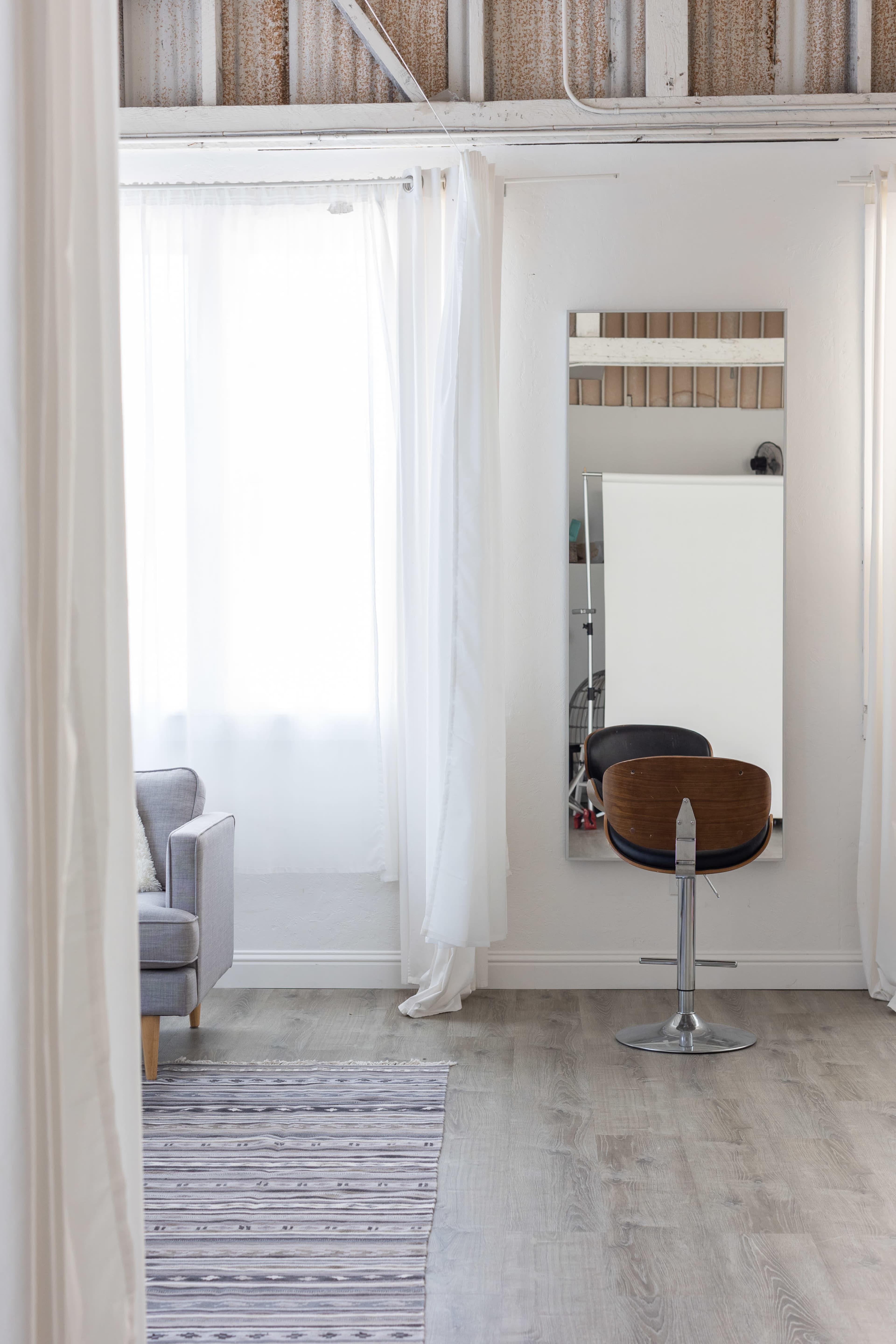 A bright, minimalistic room featuring a gray sofa, a patterned rug, and a mirror with a chair in front of it.