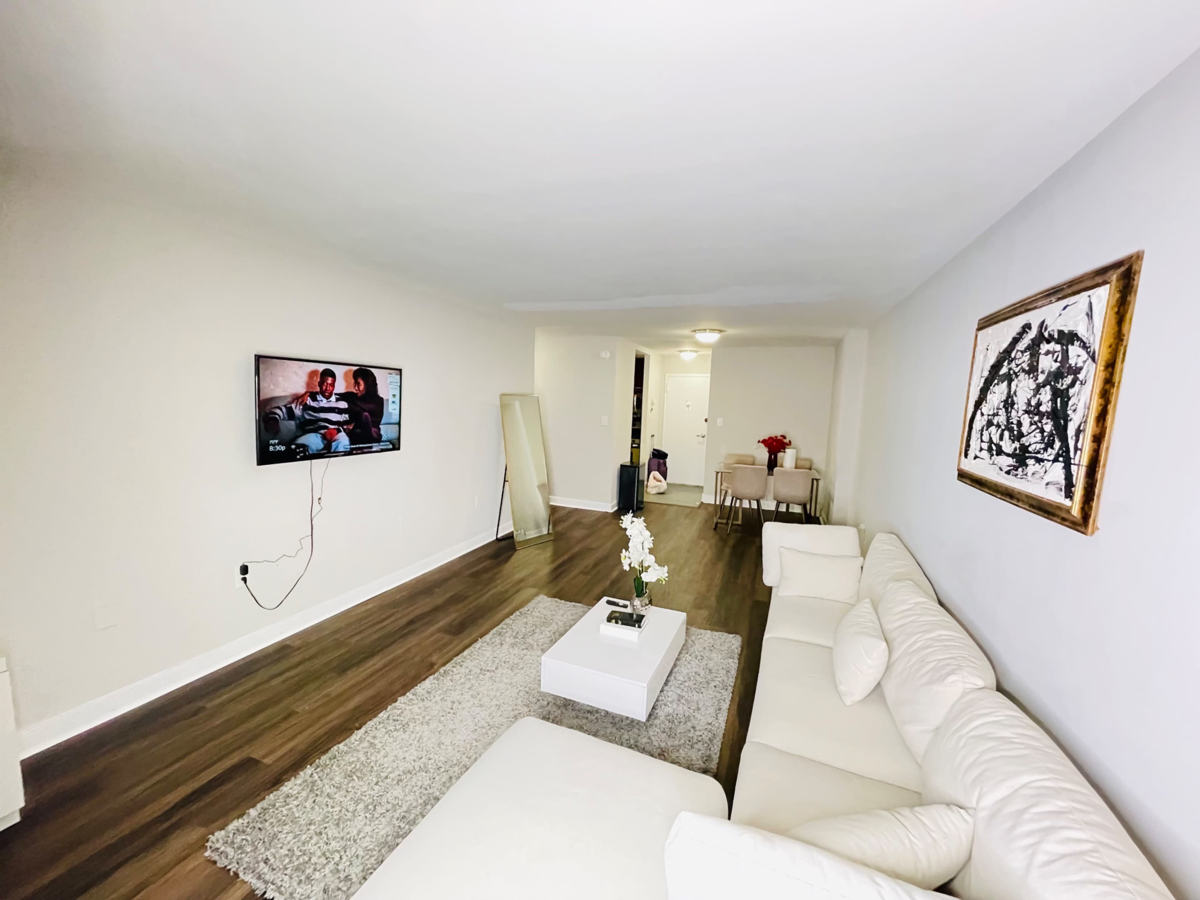 A modern living room features a white sectional sofa, a coffee table on a gray rug, a television mounted on the wall, and a dining area in the background.