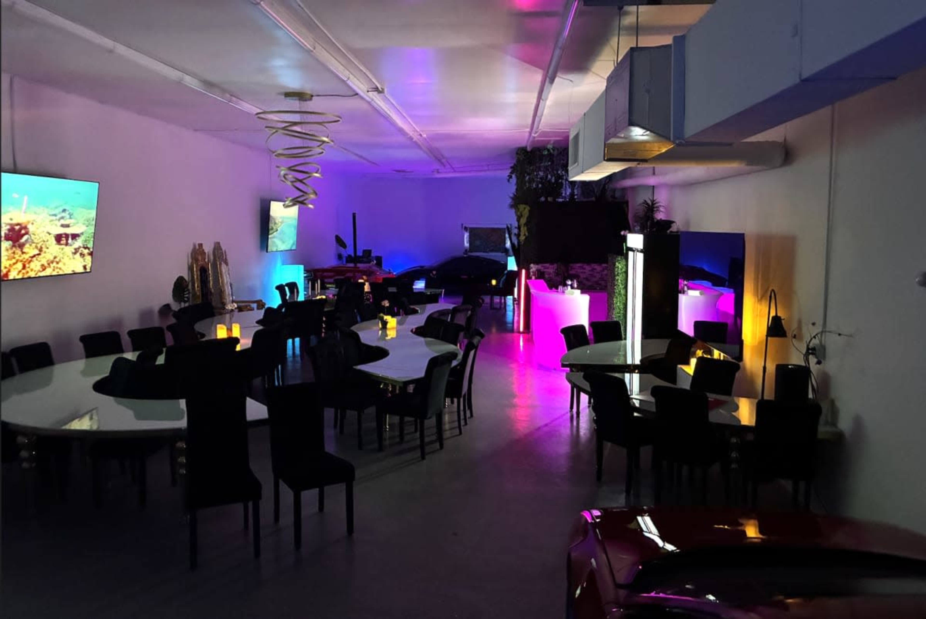 The image shows a spacious room with a large circular table surrounded by black chairs, illuminated by colorful lighting and featuring several screens on the walls.
