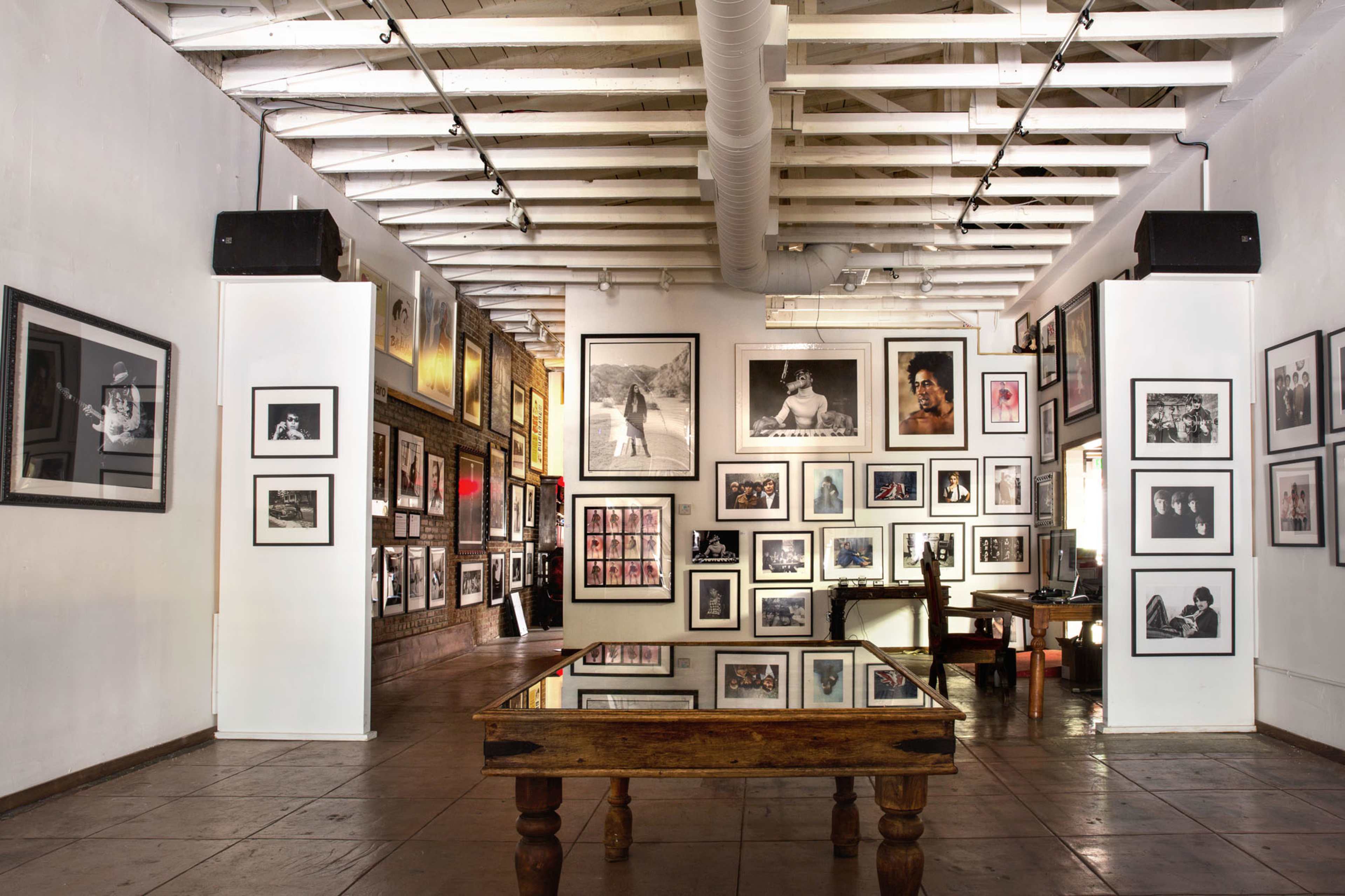 An art gallery interior with numerous framed black-and-white photographs displayed on white and brick walls, along with a wooden table in the center.