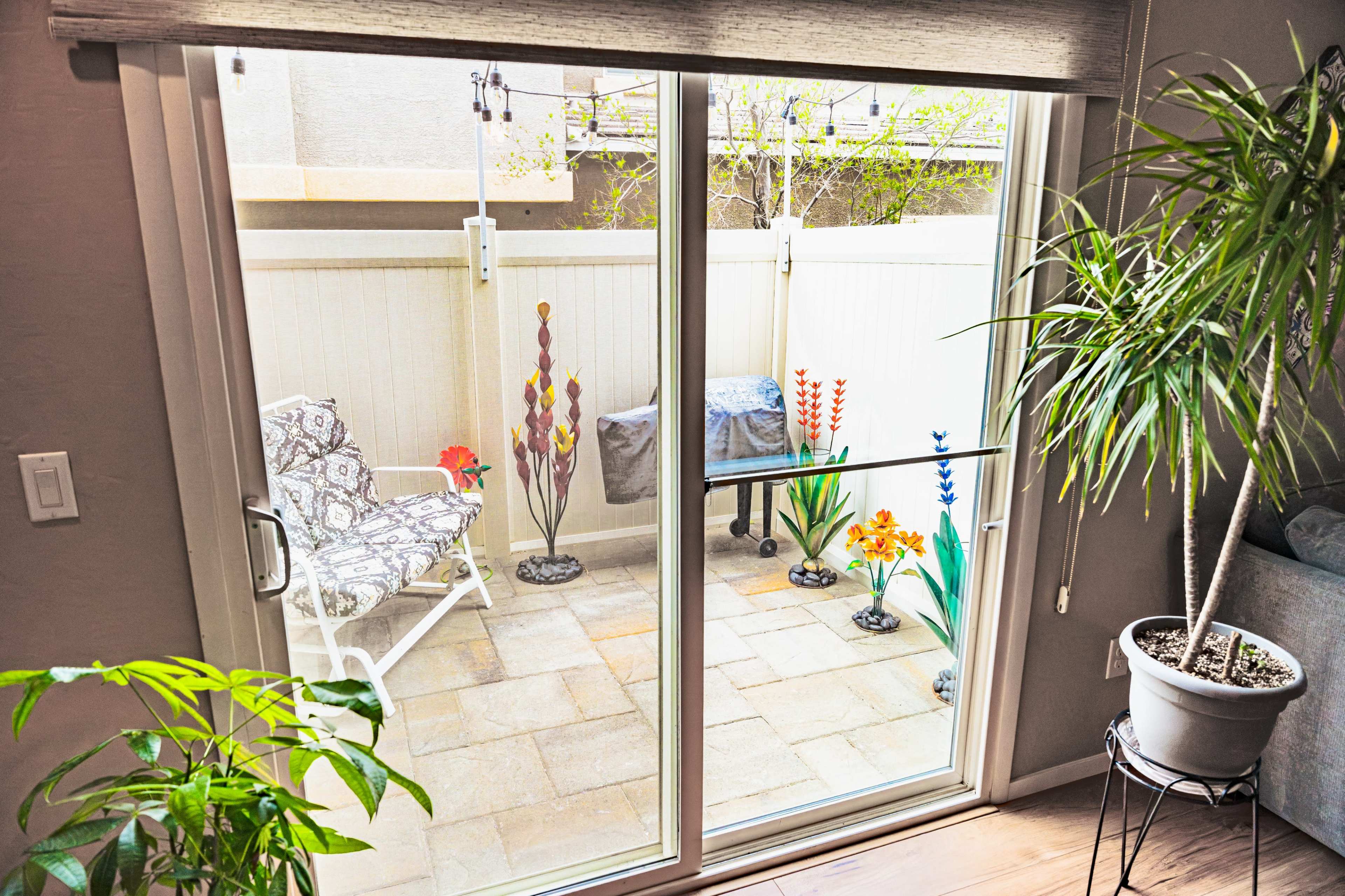 The image shows a glass sliding door revealing a small outdoor patio decorated with colorful flowers and a few potted plants.
