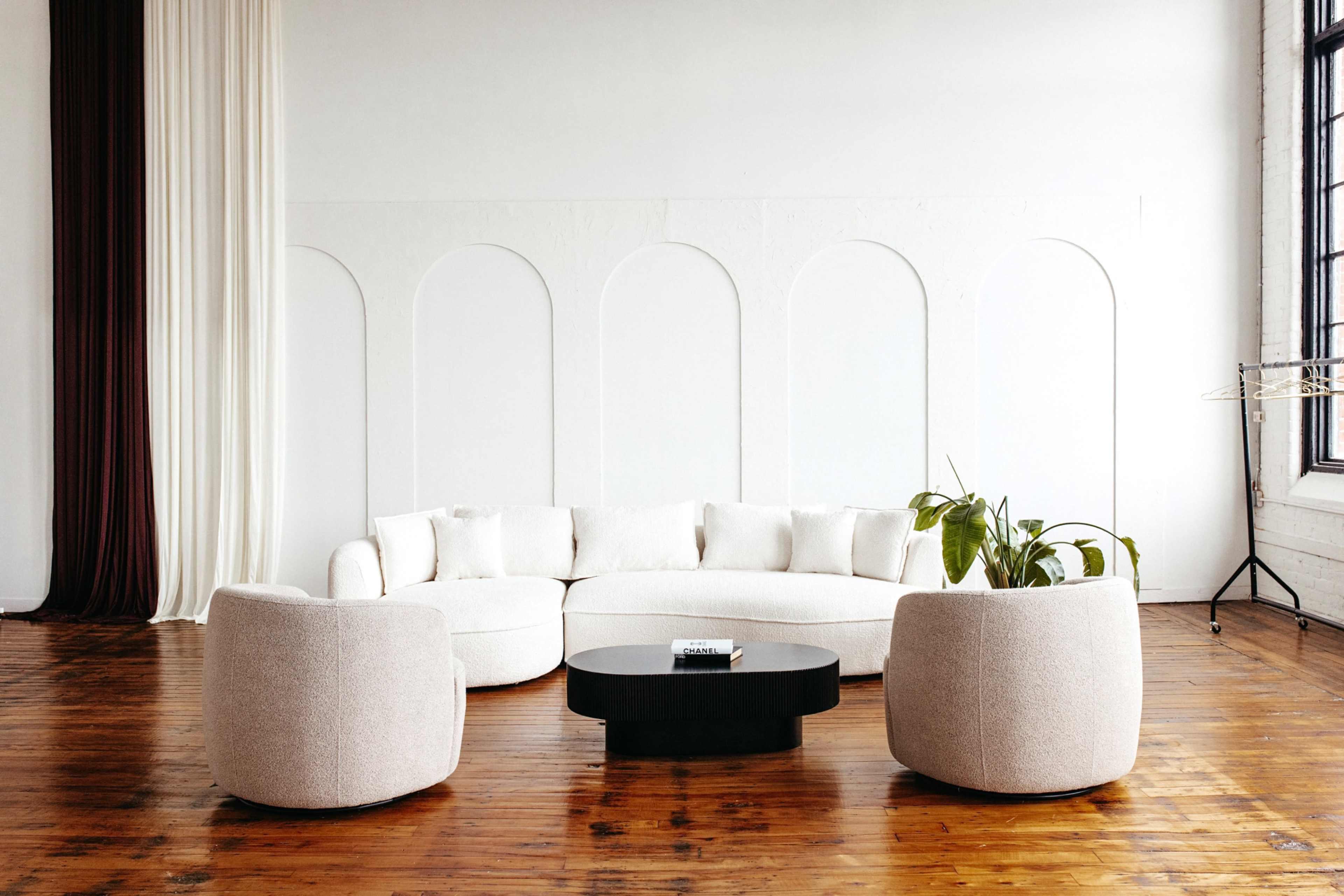 The image features a minimalist living space with a curved white sectional sofa, two round armchairs, and a black coffee table, set against a backdrop of light-colored walls and wooden floors.