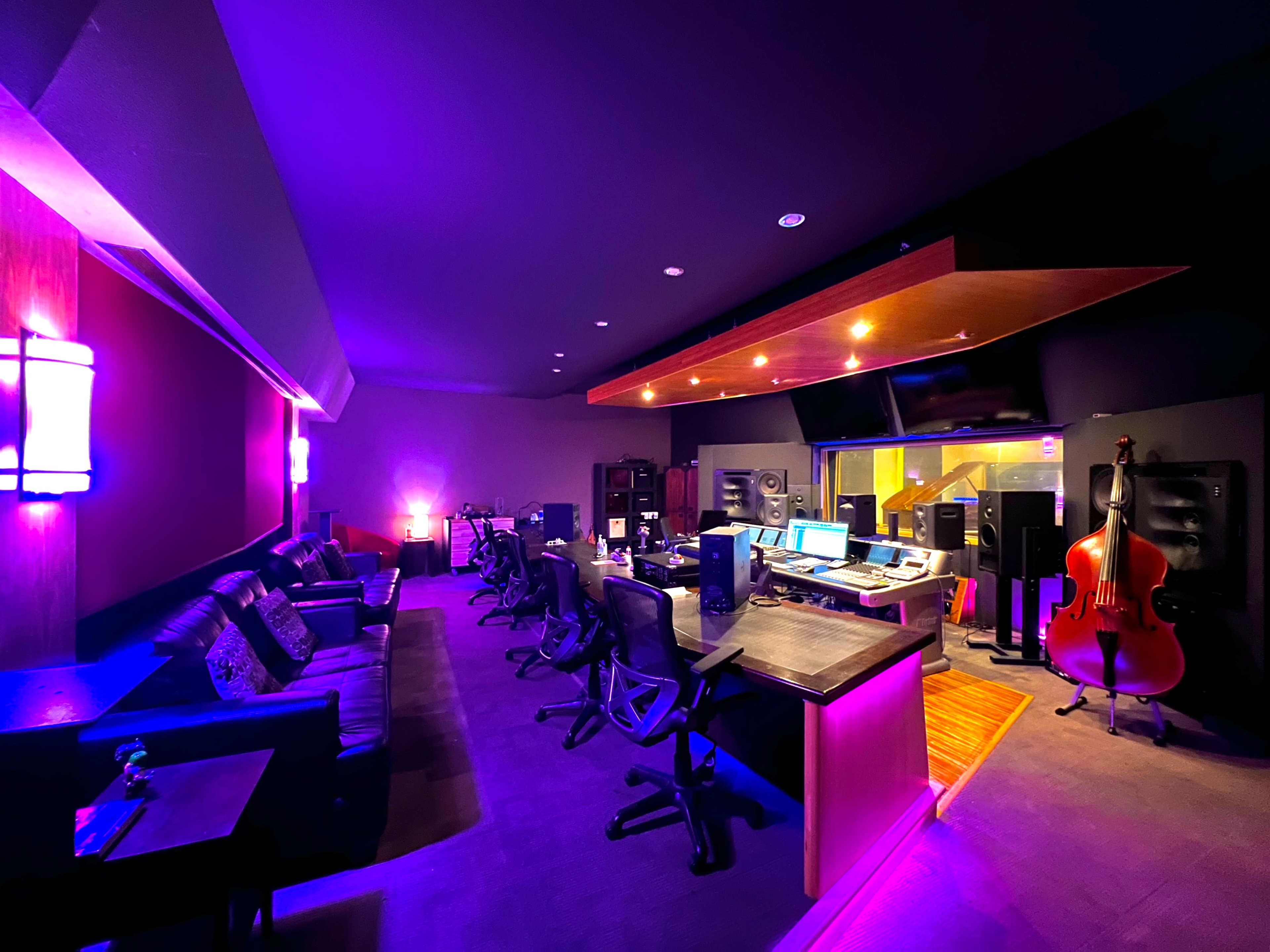 The image shows a dimly lit music studio featuring a control area with audio equipment, multiple chairs, and a large double bass in the corner.