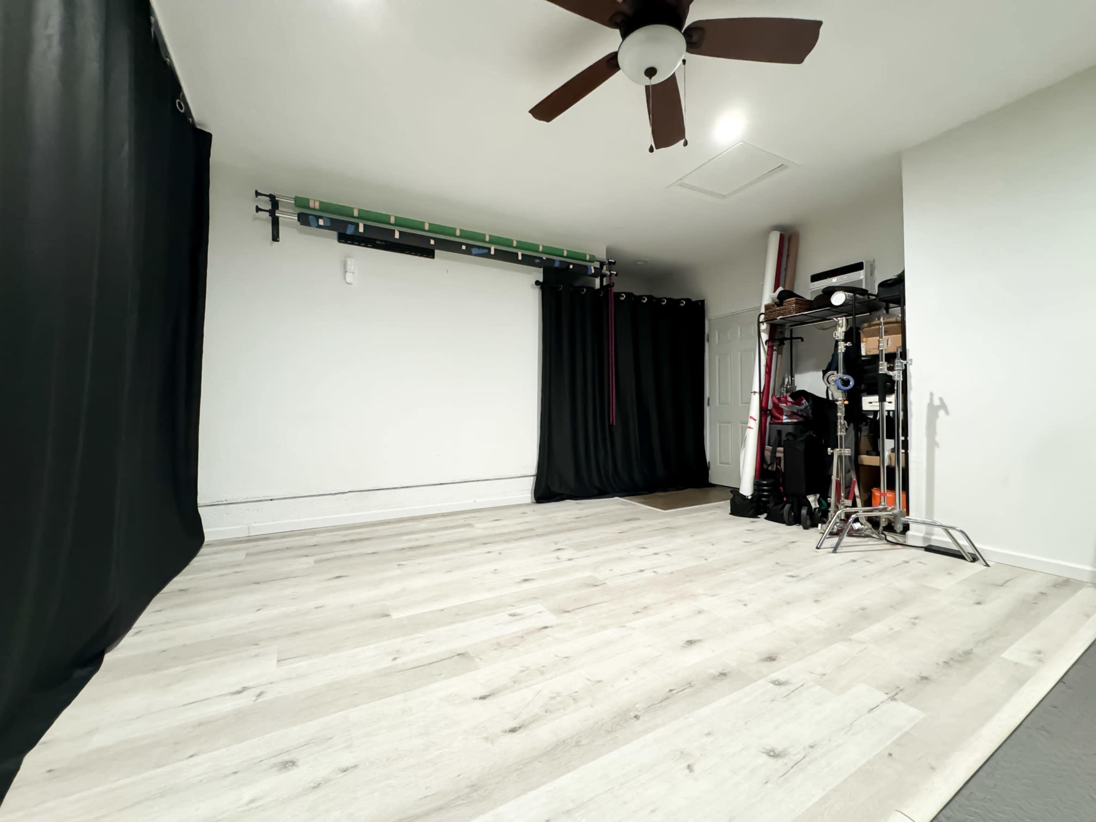 The image shows an empty room with light wood flooring, a ceiling fan, black curtains, and equipment neatly arranged against the wall.