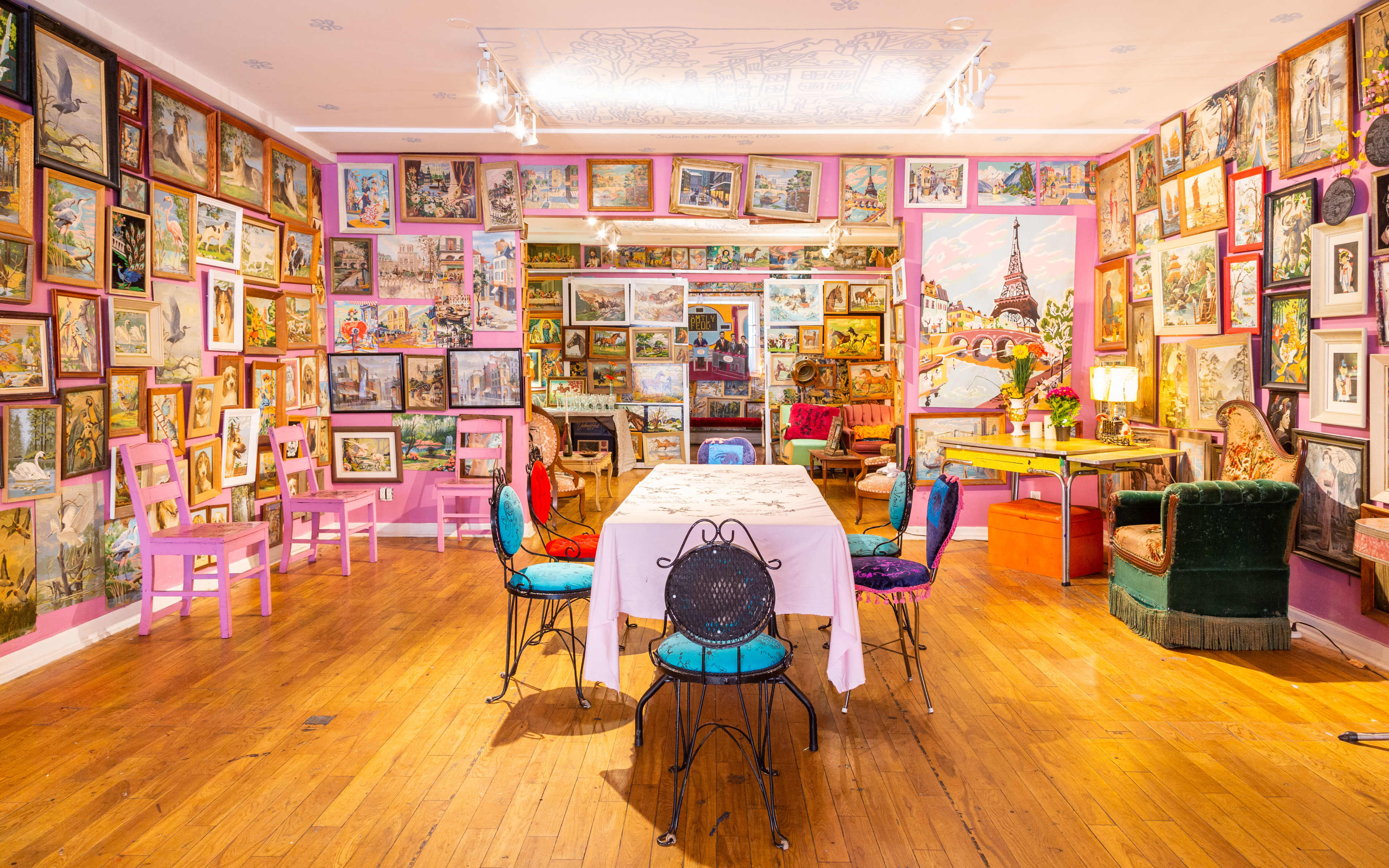 The room features a brightly decorated interior with colorful chairs around a central table, surrounded by walls adorned with numerous framed artworks.