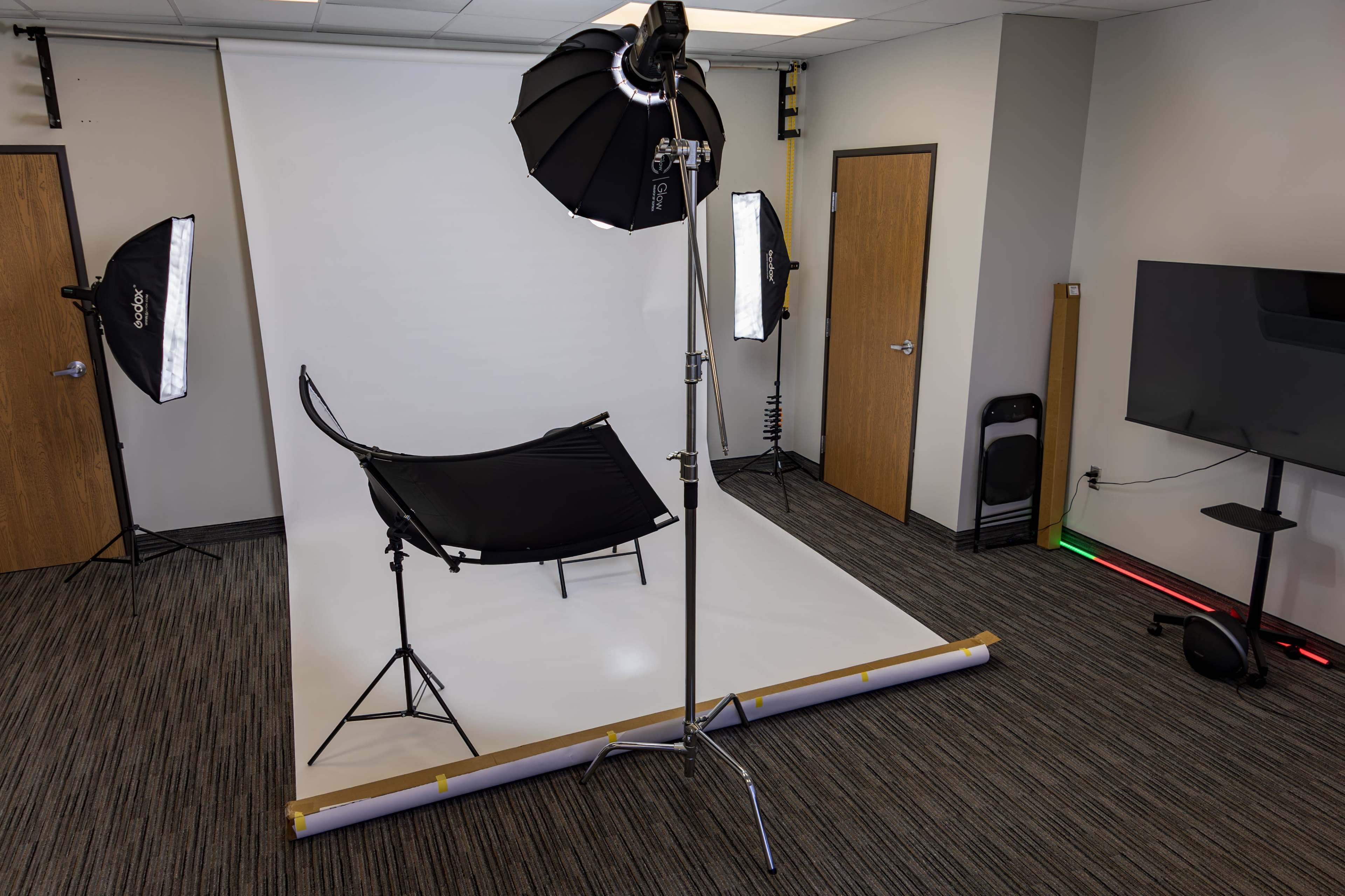 The image shows a photography studio setup with a white backdrop, lighting equipment, and a black chair positioned in front.