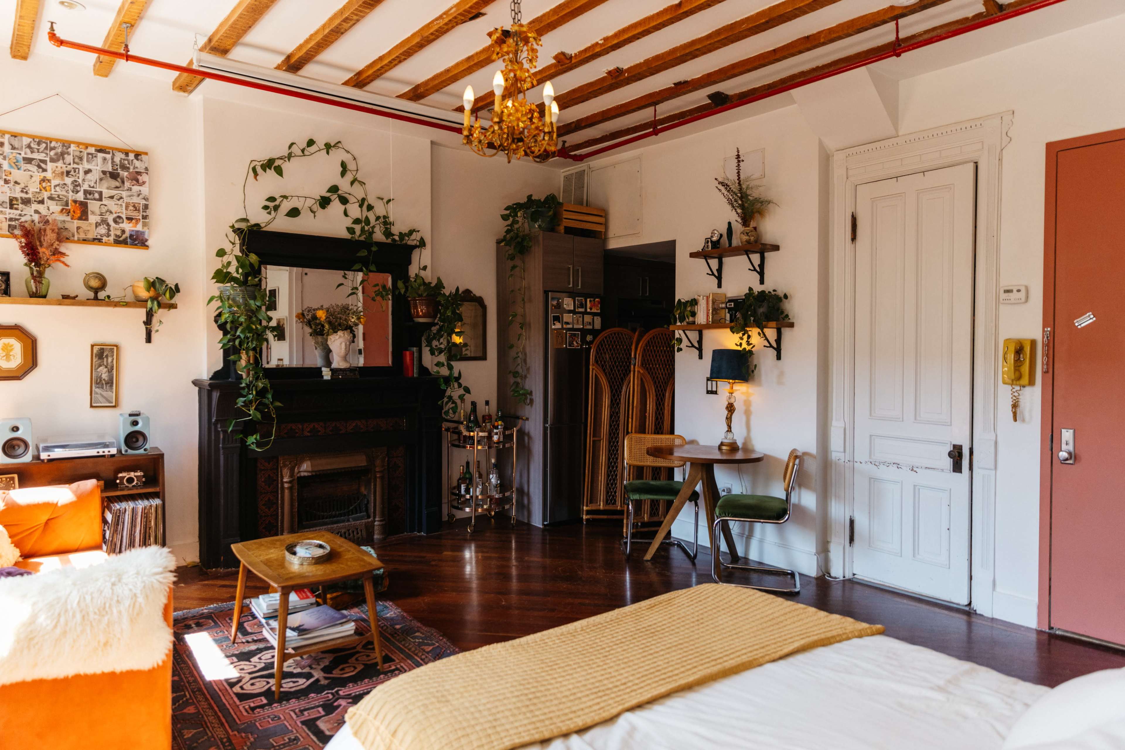 A cozy room featuring a vintage fireplace, a mix of plants adorning the walls, and various pieces of furniture including a bed, a wooden table, and a green chair.