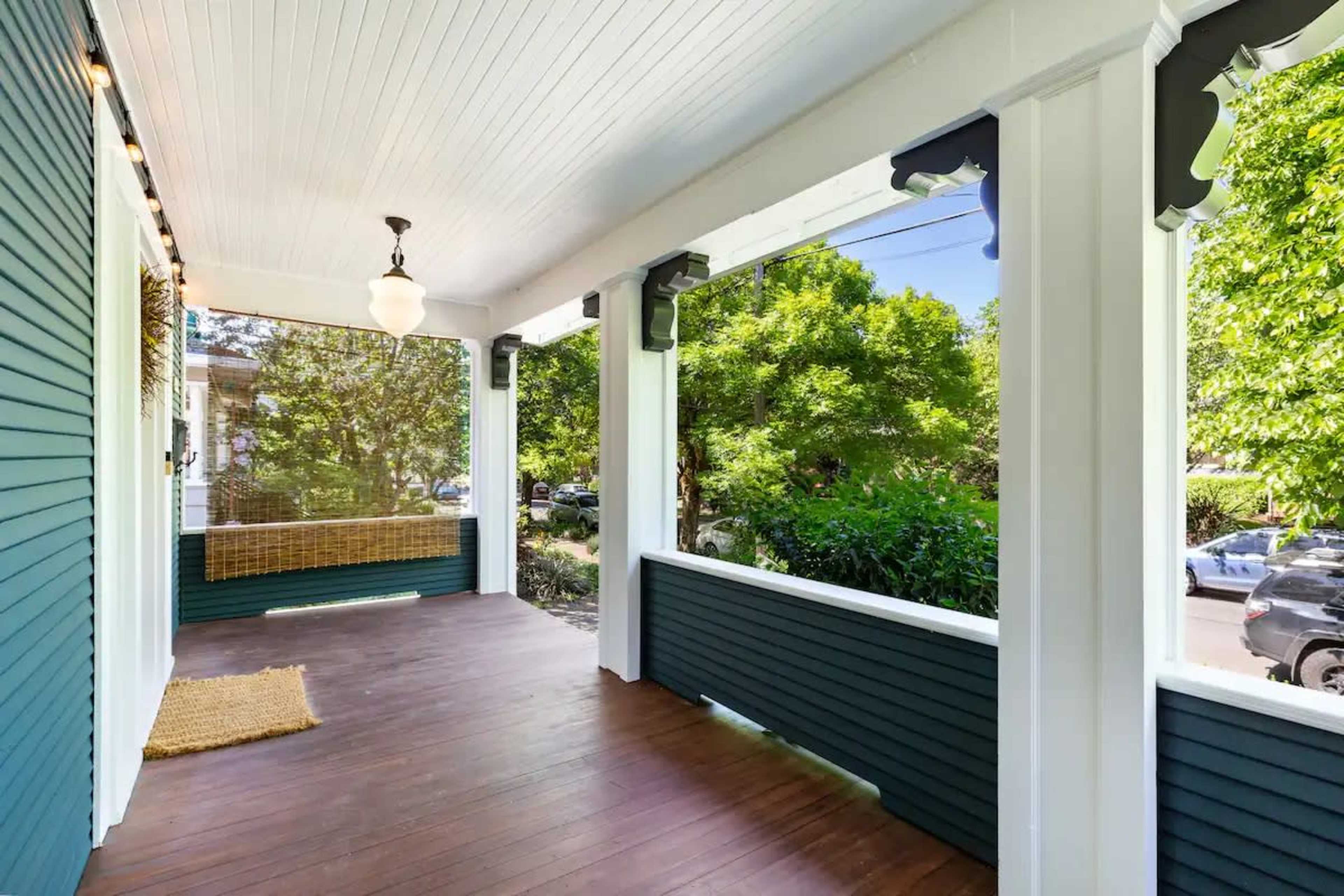 A covered porch with wooden flooring and white pillars, surrounded by greenery visible through open railings.