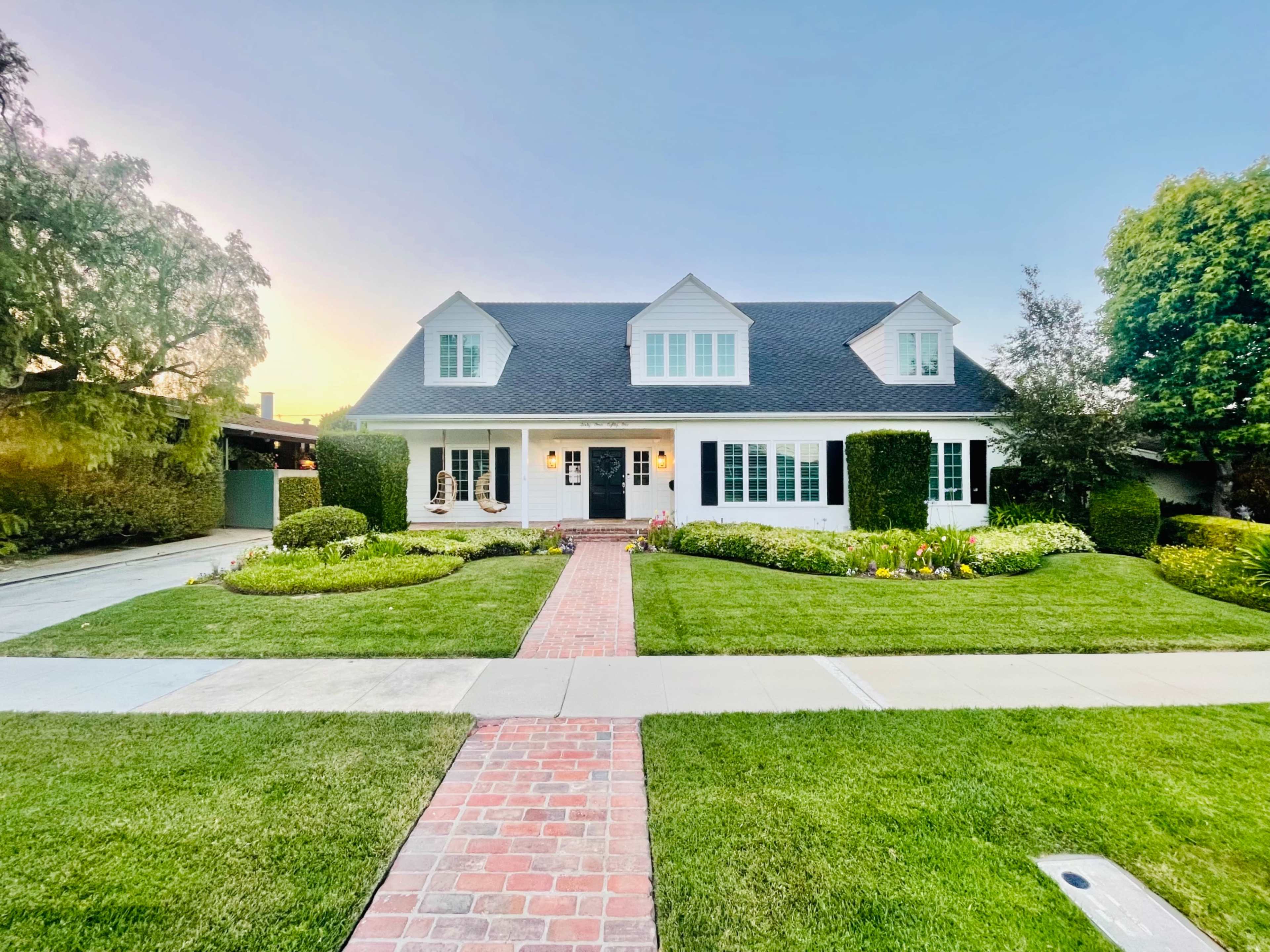 A two-story white house with a dark roof and neatly trimmed hedges is surrounded by a manicured lawn and colorful flower beds.