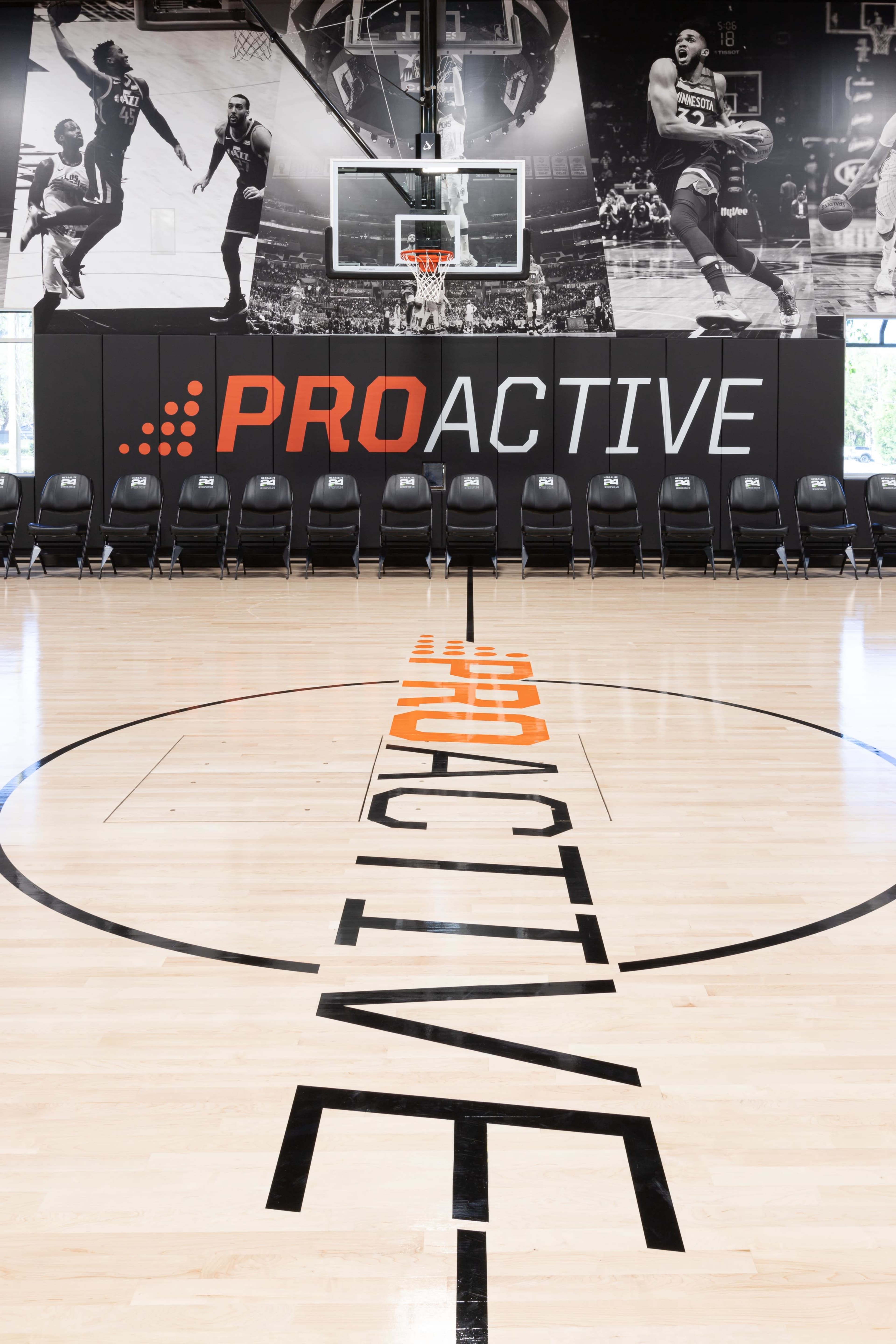 The image shows a basketball court with a prominent "PROACTIVE" logo at center court and bleachers along one side featuring sports graphics on the wall behind the hoop.