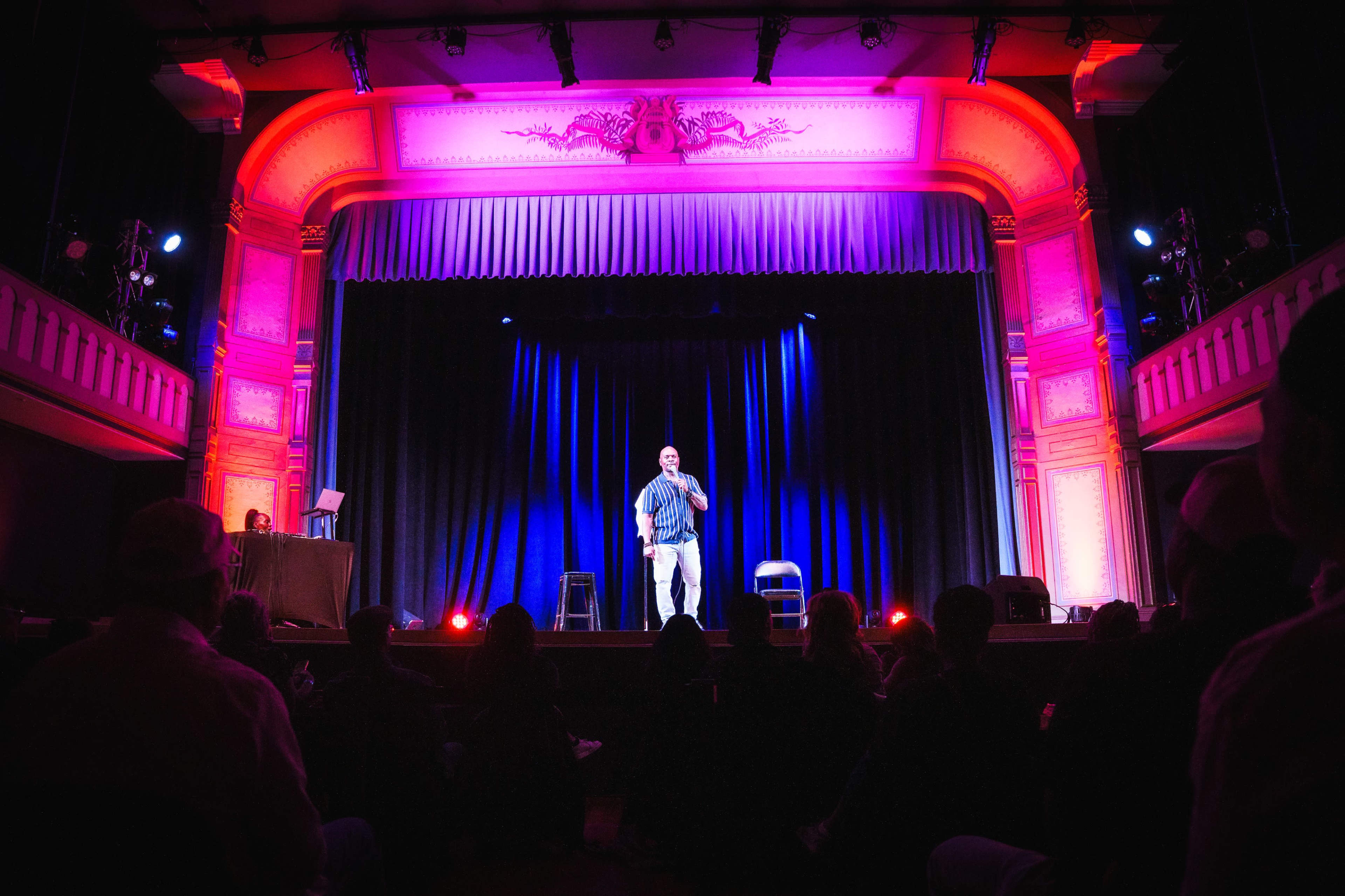 A comedian performs on stage in a brightly lit theater, with a dark curtain backdrop and an audience seated in shadow.