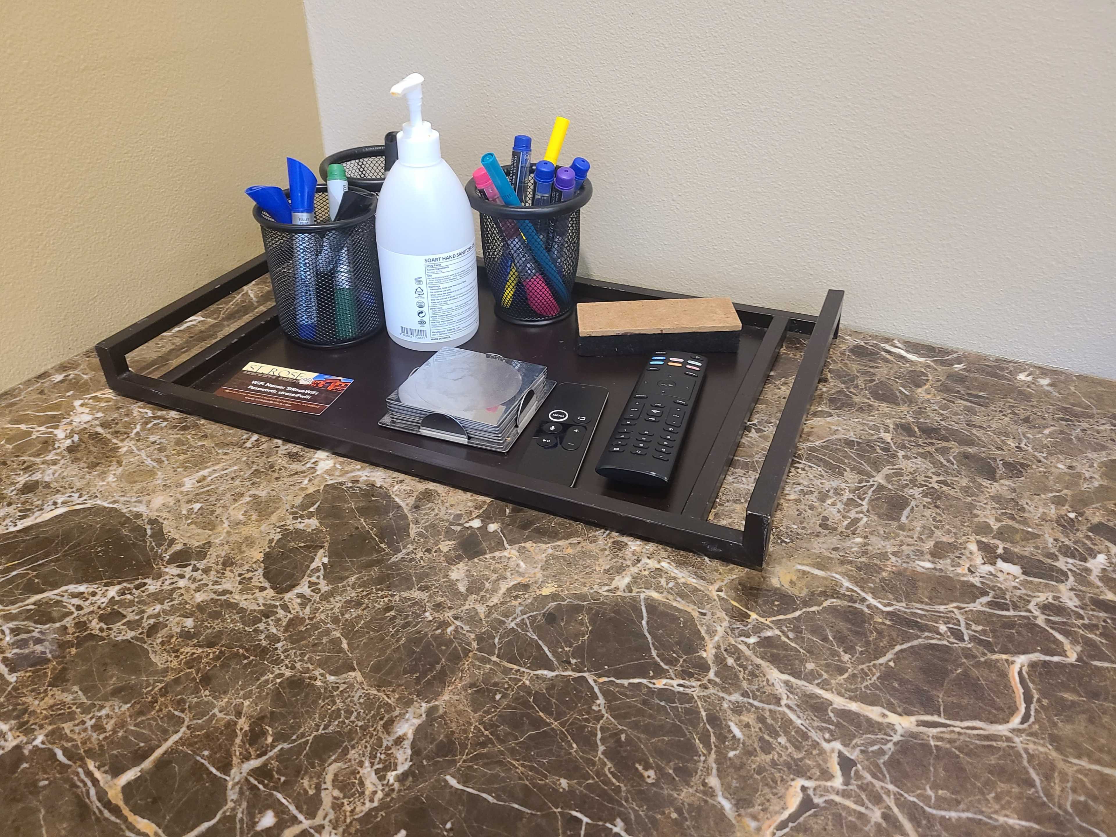 A rectangular tray rests on a marble countertop, holding a hand sanitizer, two pen holders filled with markers and pens, a remote control, a stone, and a stack of coasters.