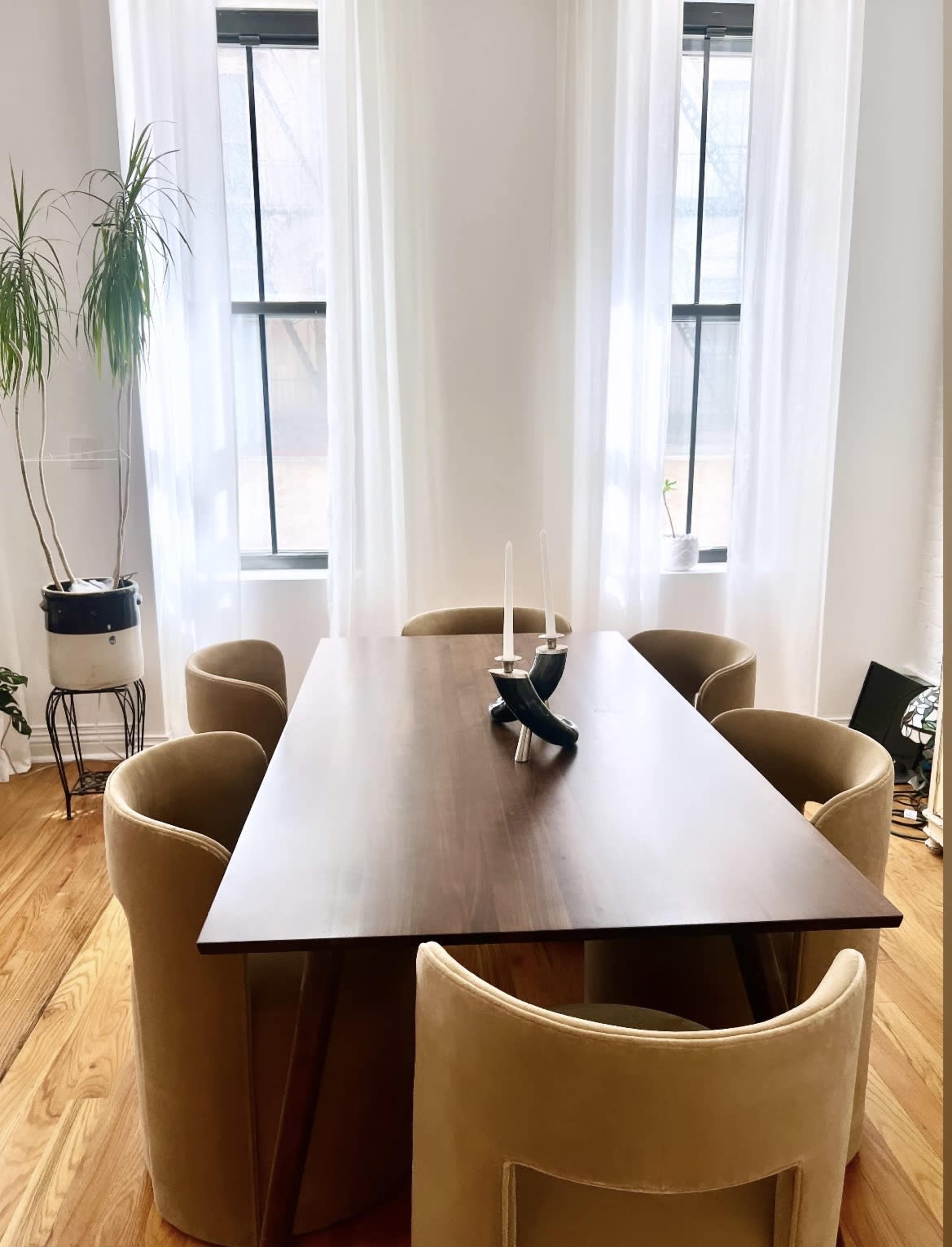 A wooden dining table is surrounded by four beige chairs under windows adorned with sheer white curtains.