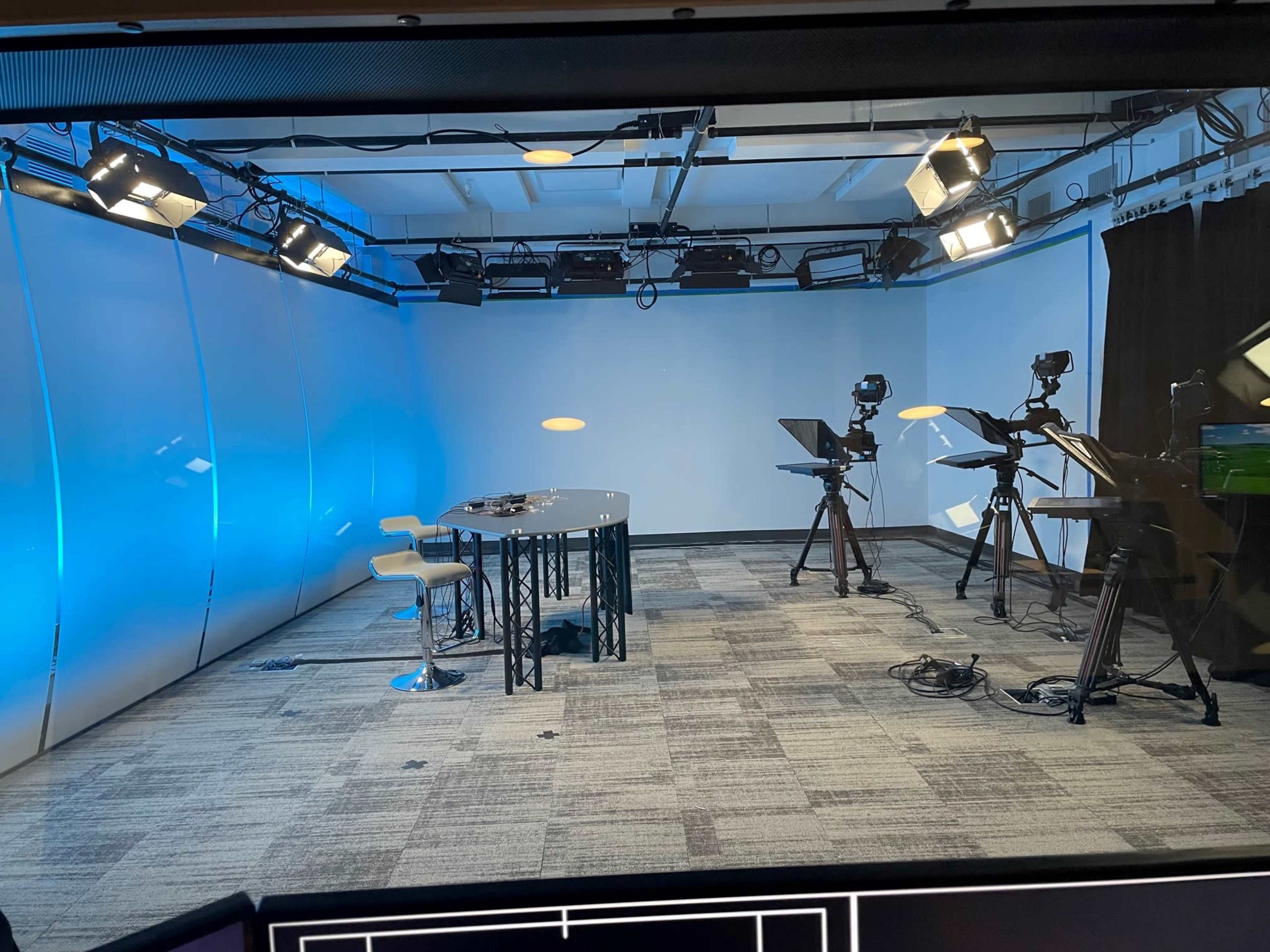 A well-lit television studio with a central table and three camera setups positioned on the side.