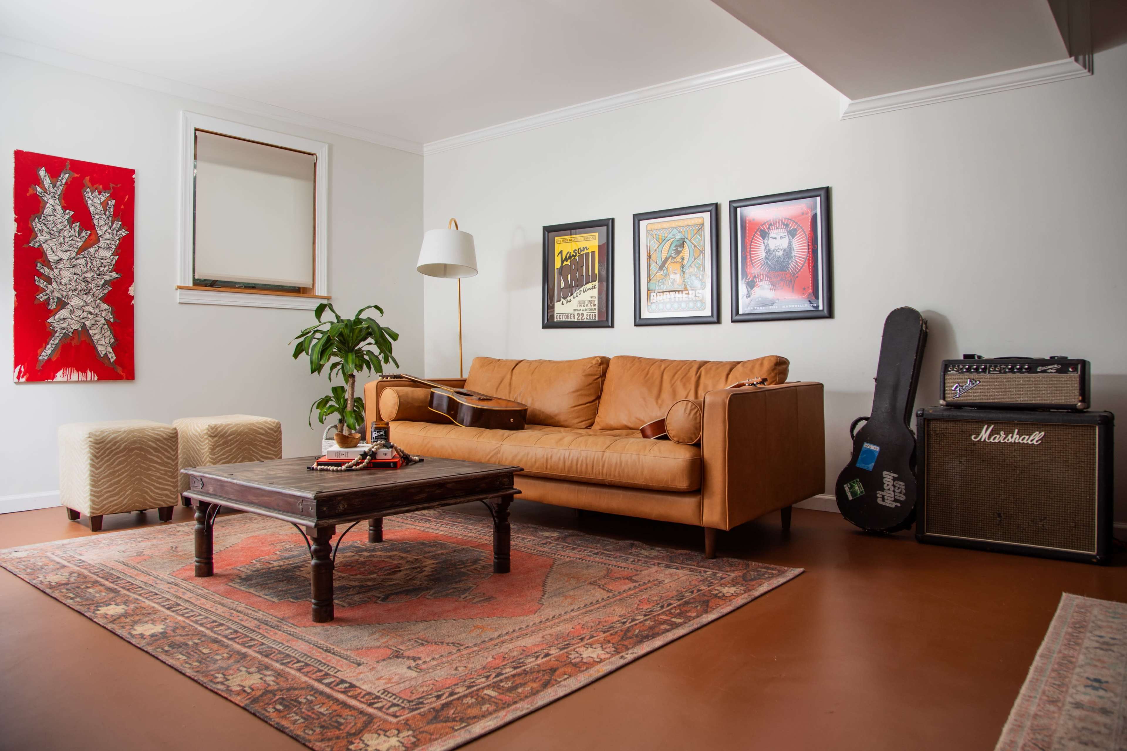 A cozy living room features a brown leather couch, a coffee table, framed artwork on the walls, a guitar resting on the couch, and an amplifier in the corner.