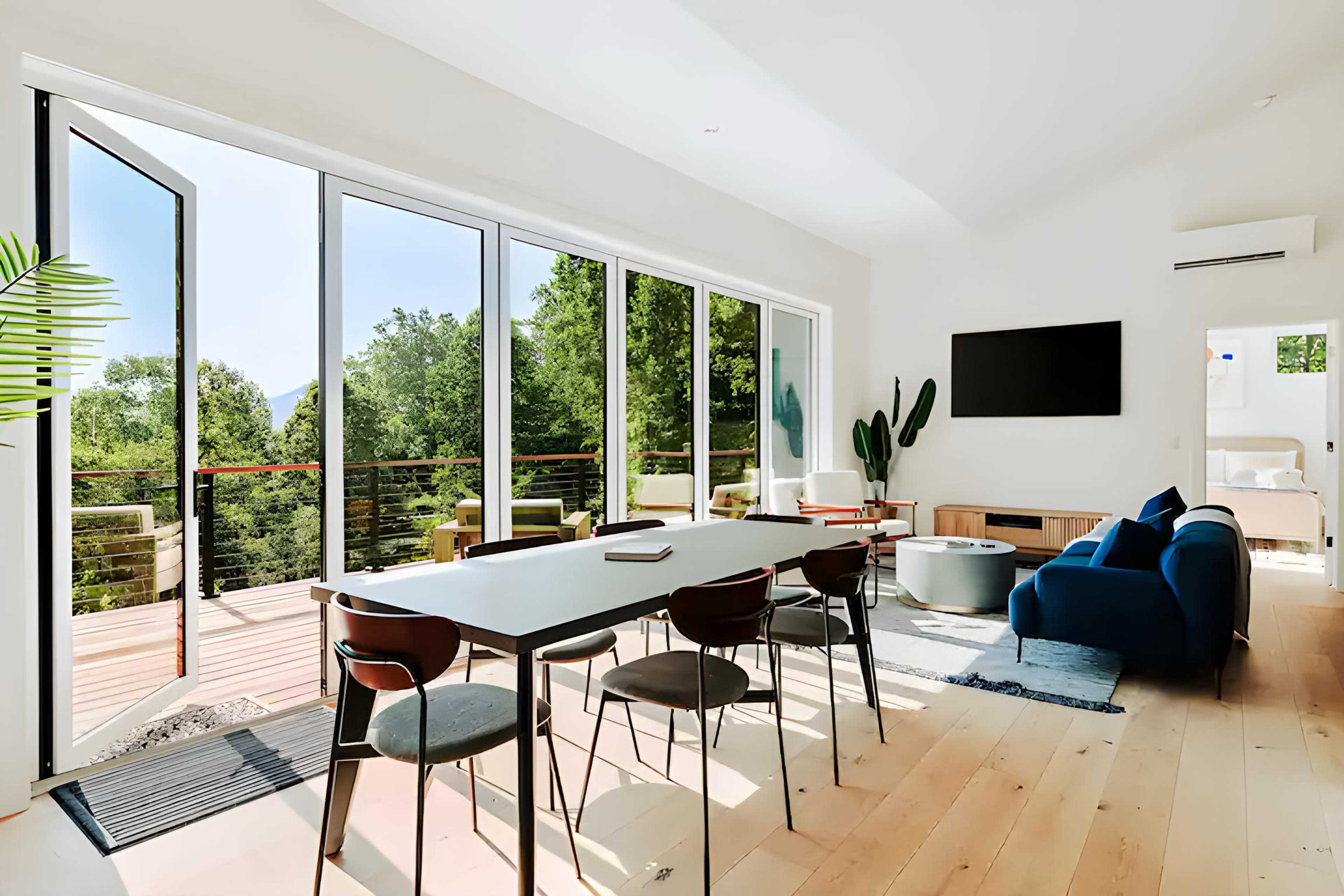 A modern living space features a large dining table, sliding glass doors leading to a balcony with trees outside, and a blue sofa alongside a wall-mounted television.