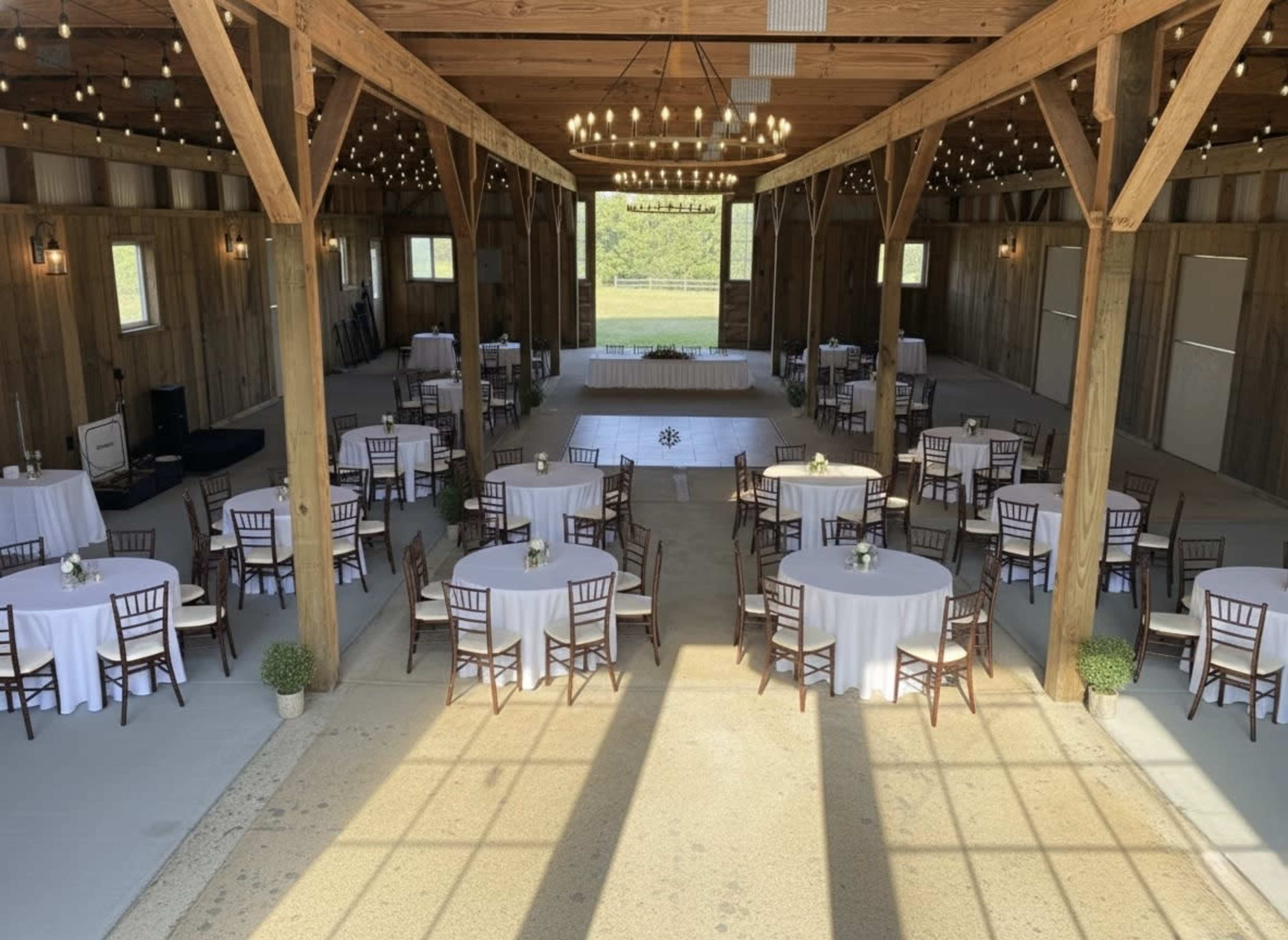 The image shows a spacious venue with wooden beams, several round tables set with white tablecloths, and a stage at one end under warm string lights.