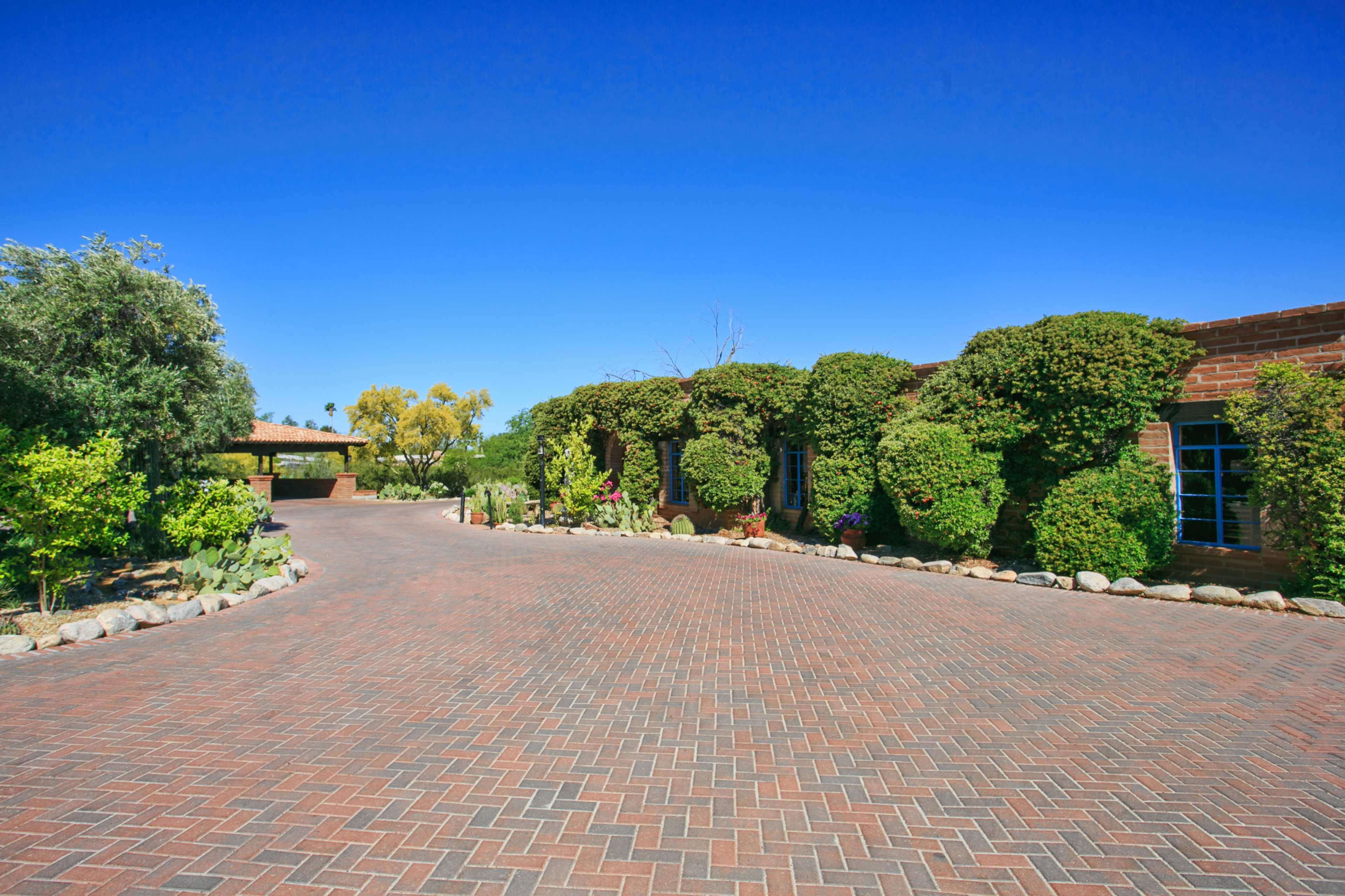 A brick-paved driveway leads to a house surrounded by well-maintained shrubs and trees under a clear blue sky.