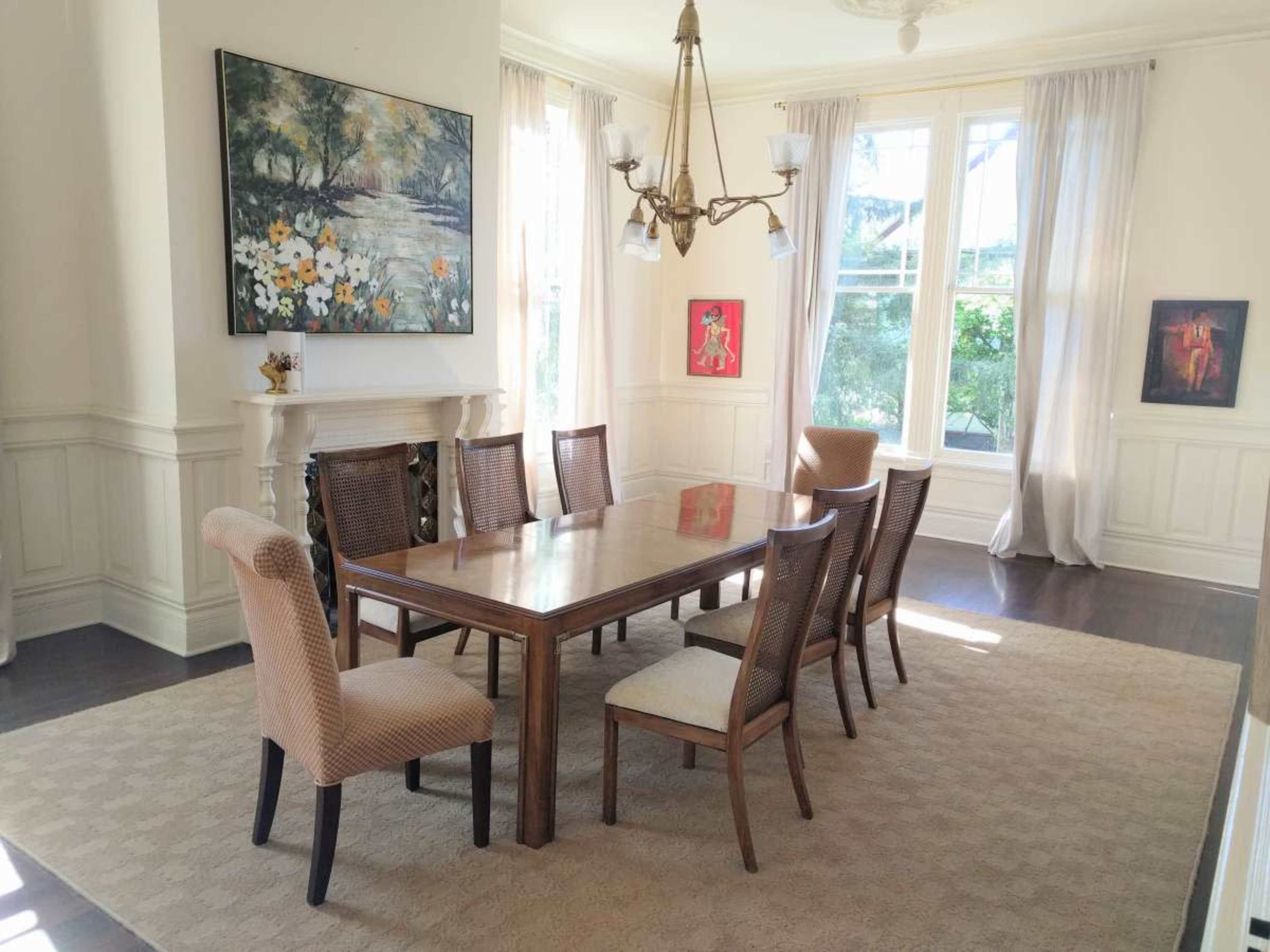 The image shows a well-lit dining room featuring a long wooden table surrounded by eight chairs, with large windows and artwork on the walls.