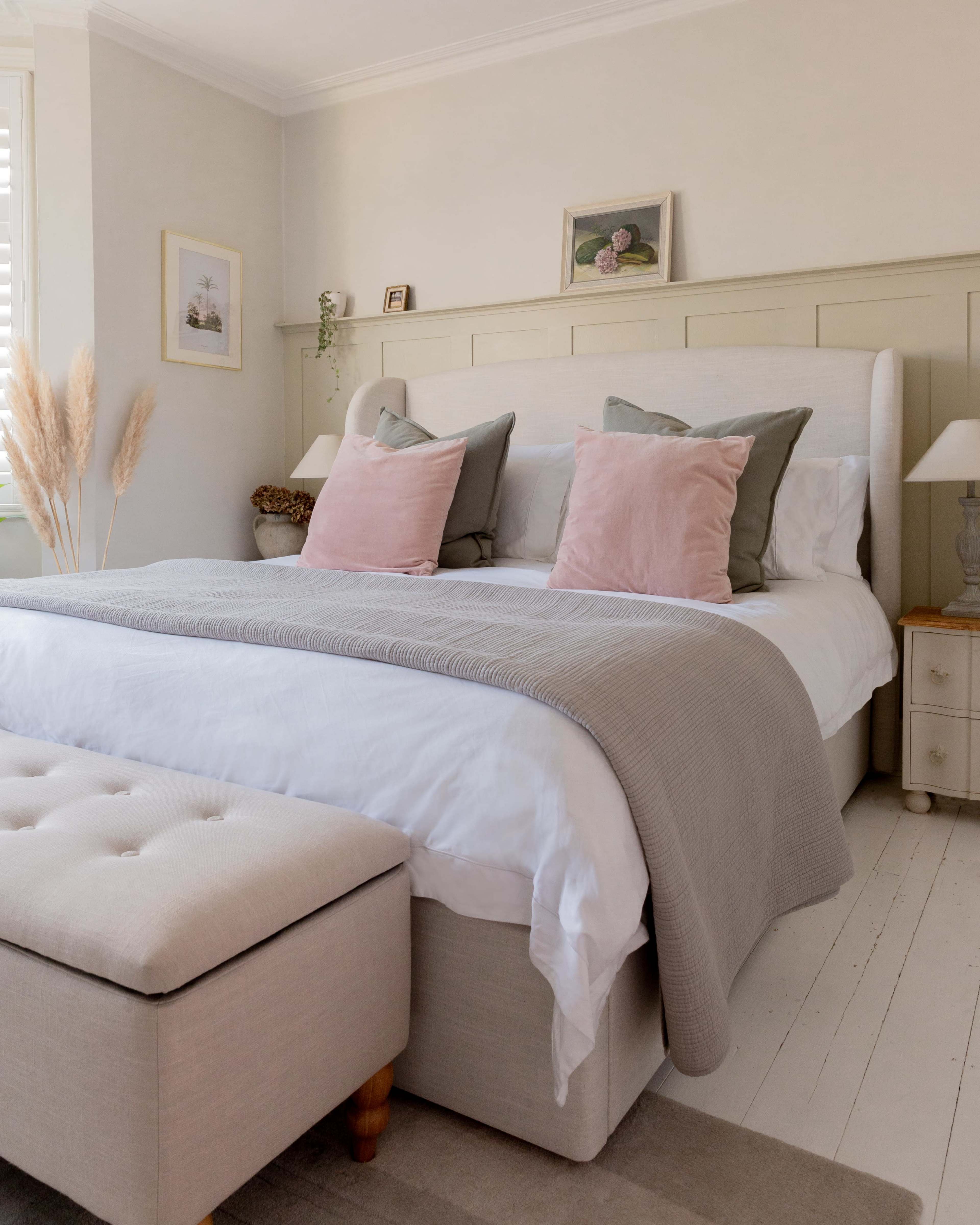 A neatly arranged bedroom features a upholstered bed with decorative pillows, a light-colored blanket, and a padded ottoman at the foot.