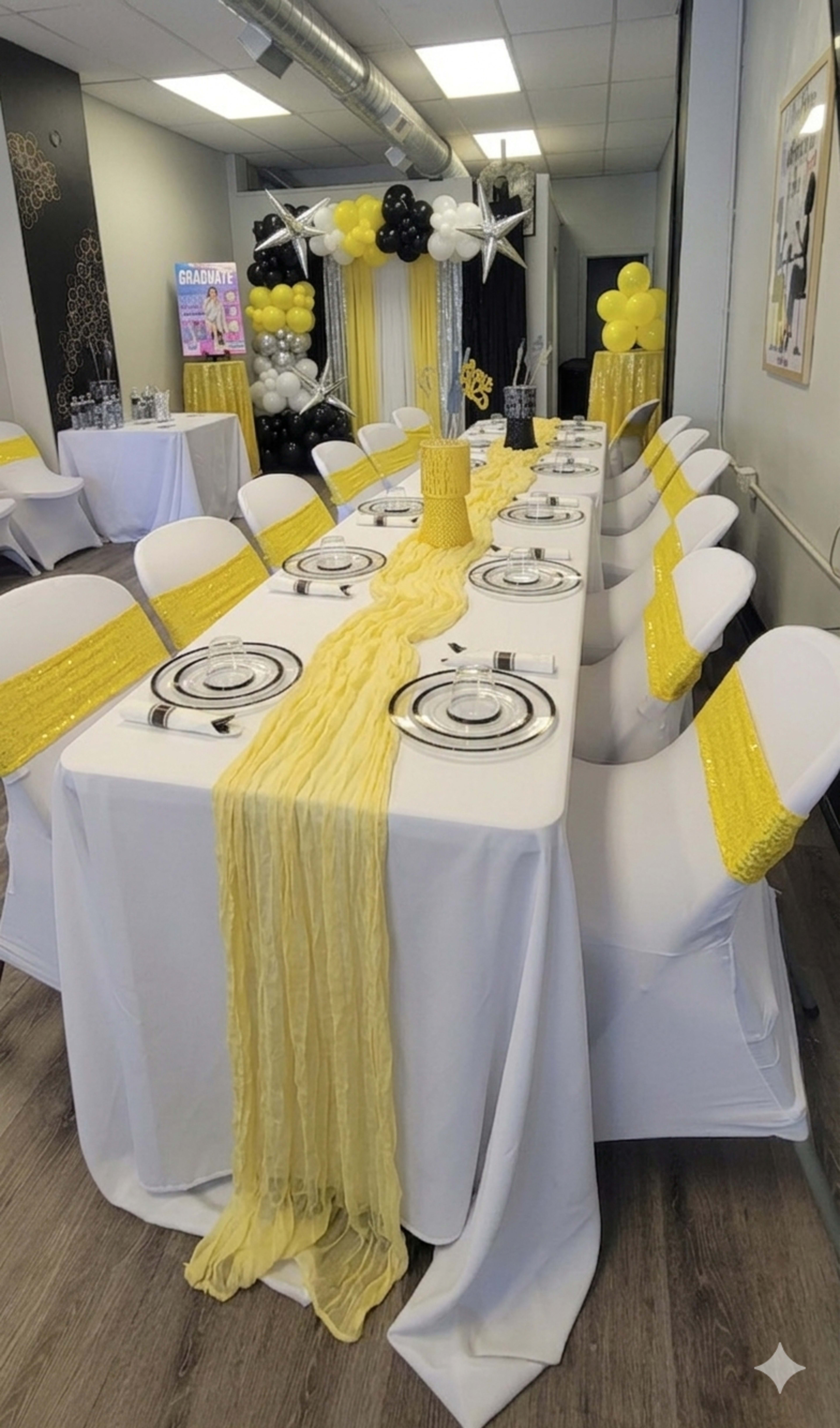 A long dining table is set with round plates and silverware, adorned with a yellow table runner and surrounded by chairs covered in white fabric with yellow sashes, in a decorated room featuring black and gold balloons.