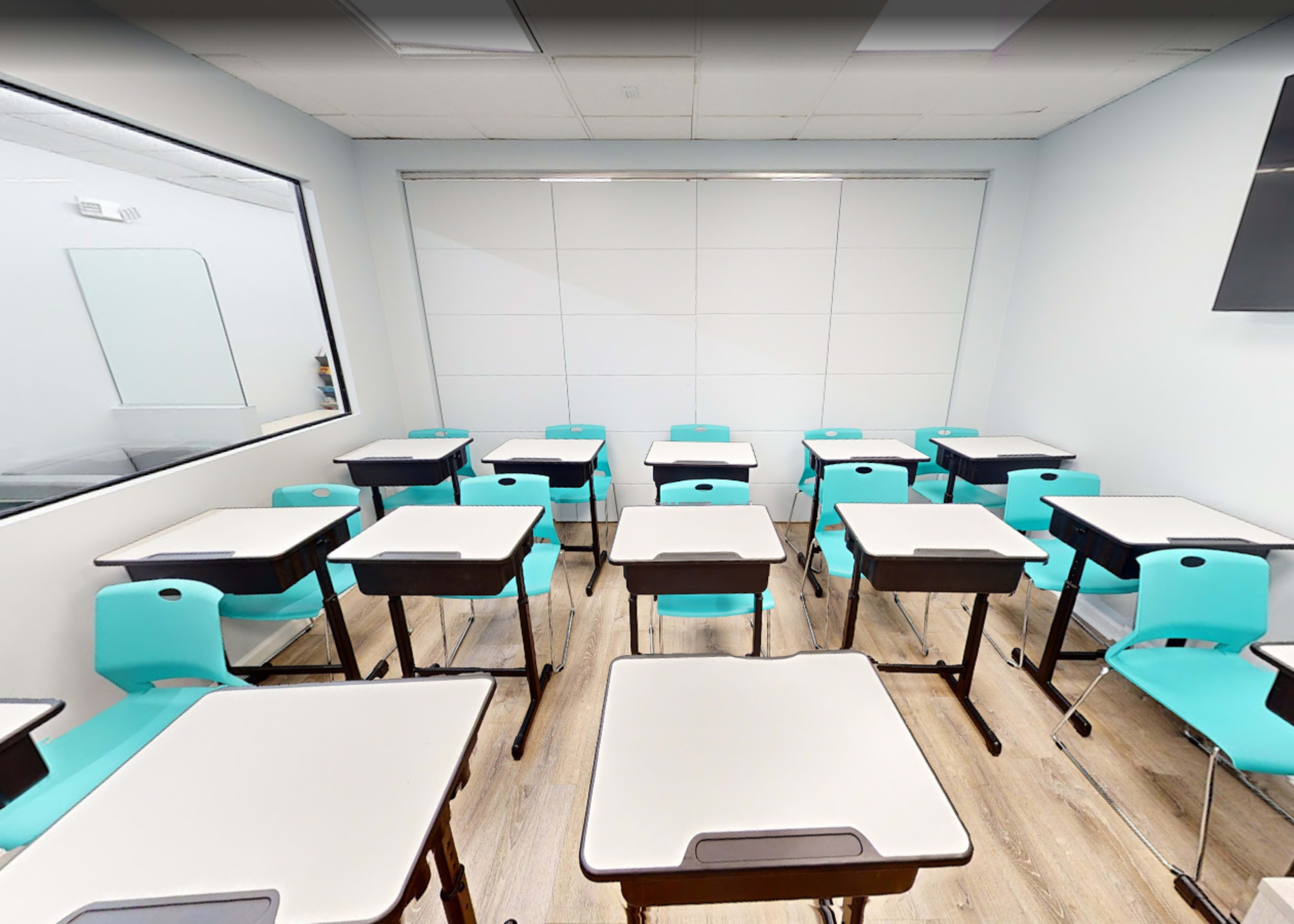 The image shows a classroom with multiple empty turquoise desks arranged facing a whiteboard.