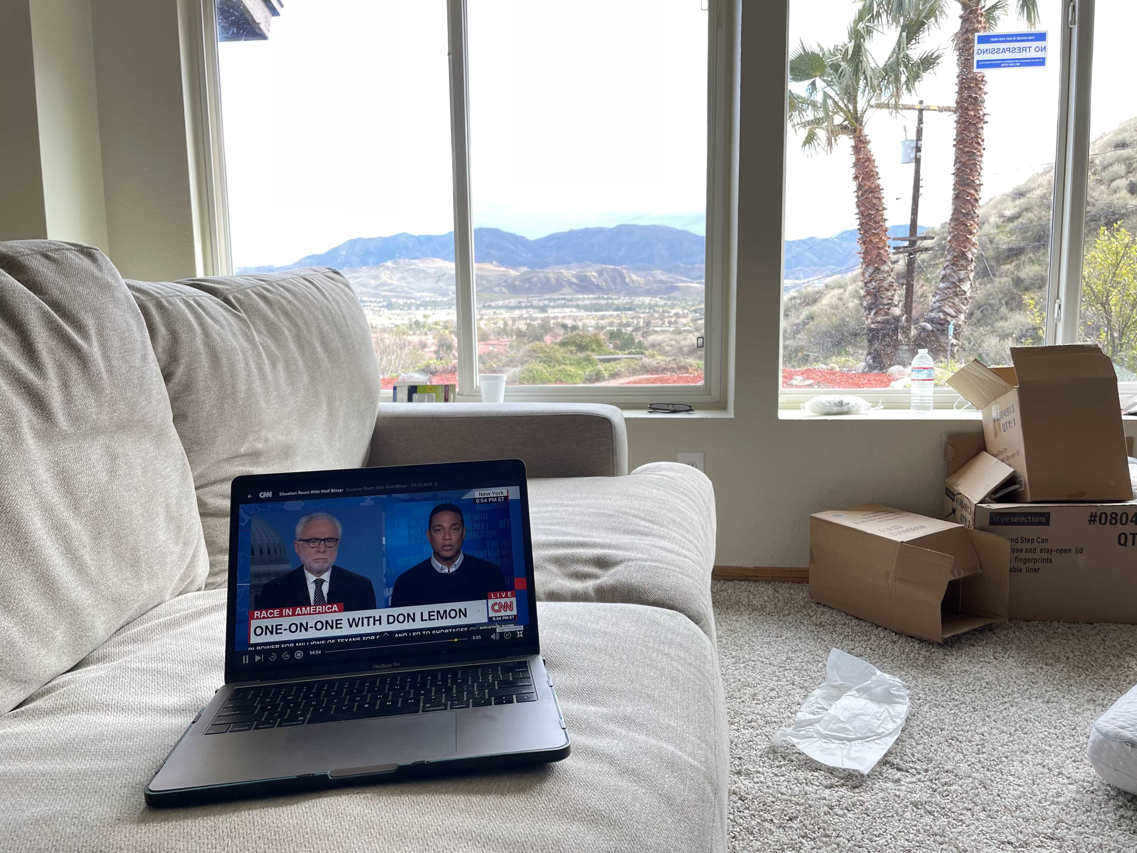 A laptop displaying a news segment is placed on a couch near a window overlooking a mountainous landscape, with unpacked boxes and crumpled paper on the floor.