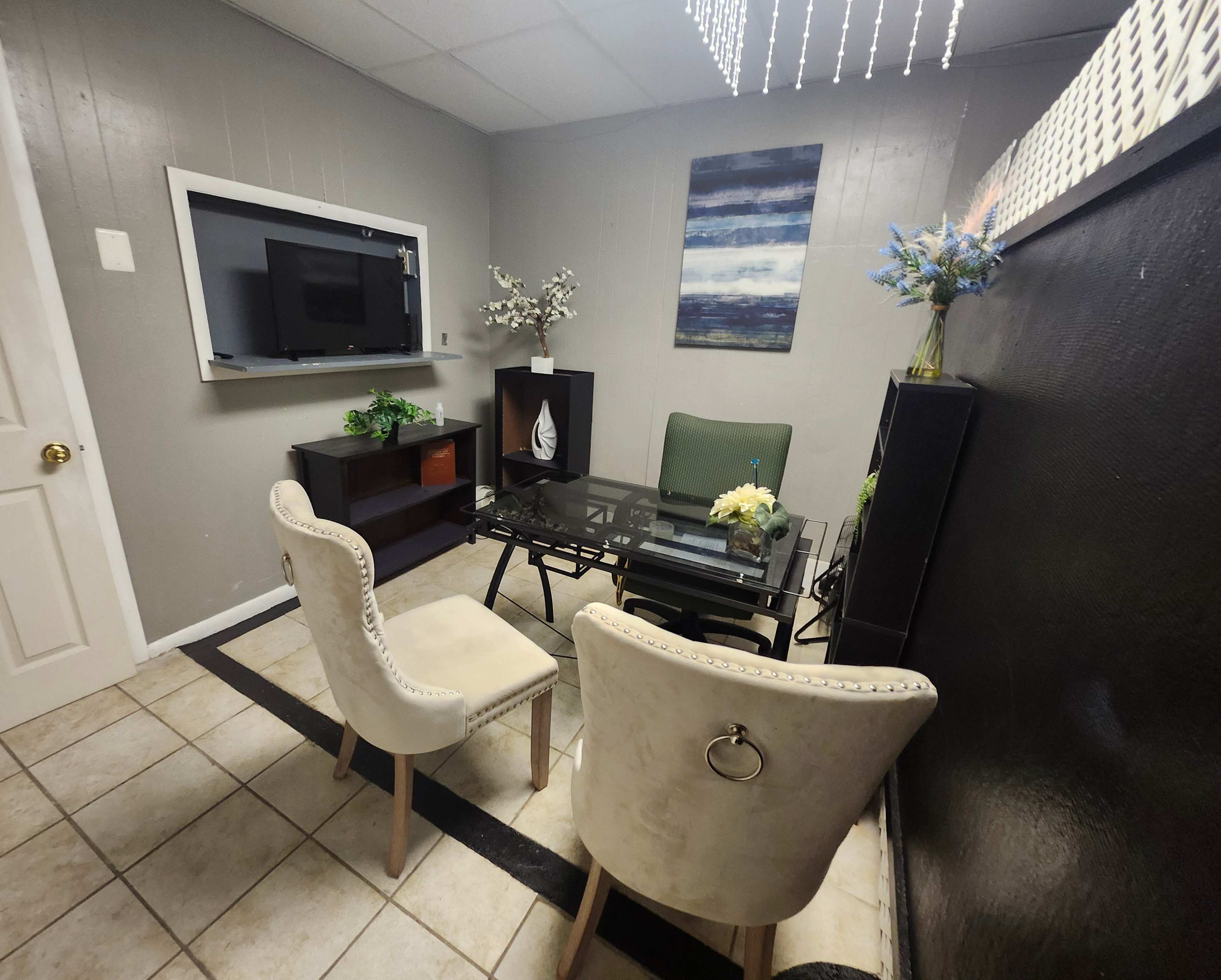 The image shows a small, well-decorated room featuring a glass table surrounded by two upholstered chairs, a wall-mounted television, and a shelf with plants and decorative items.