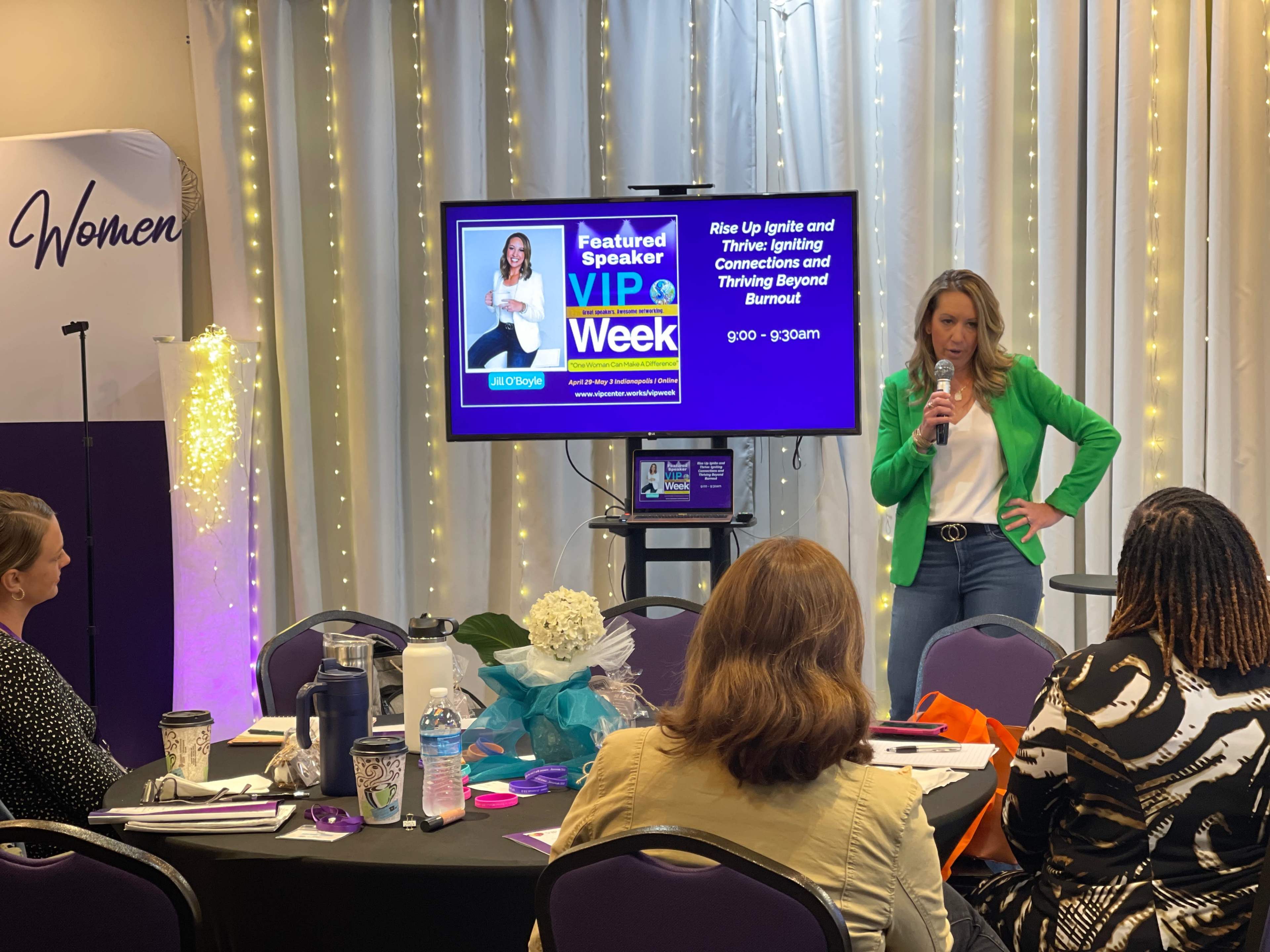 A speaker stands in front of a screen displaying event details while addressing an audience seated at tables.