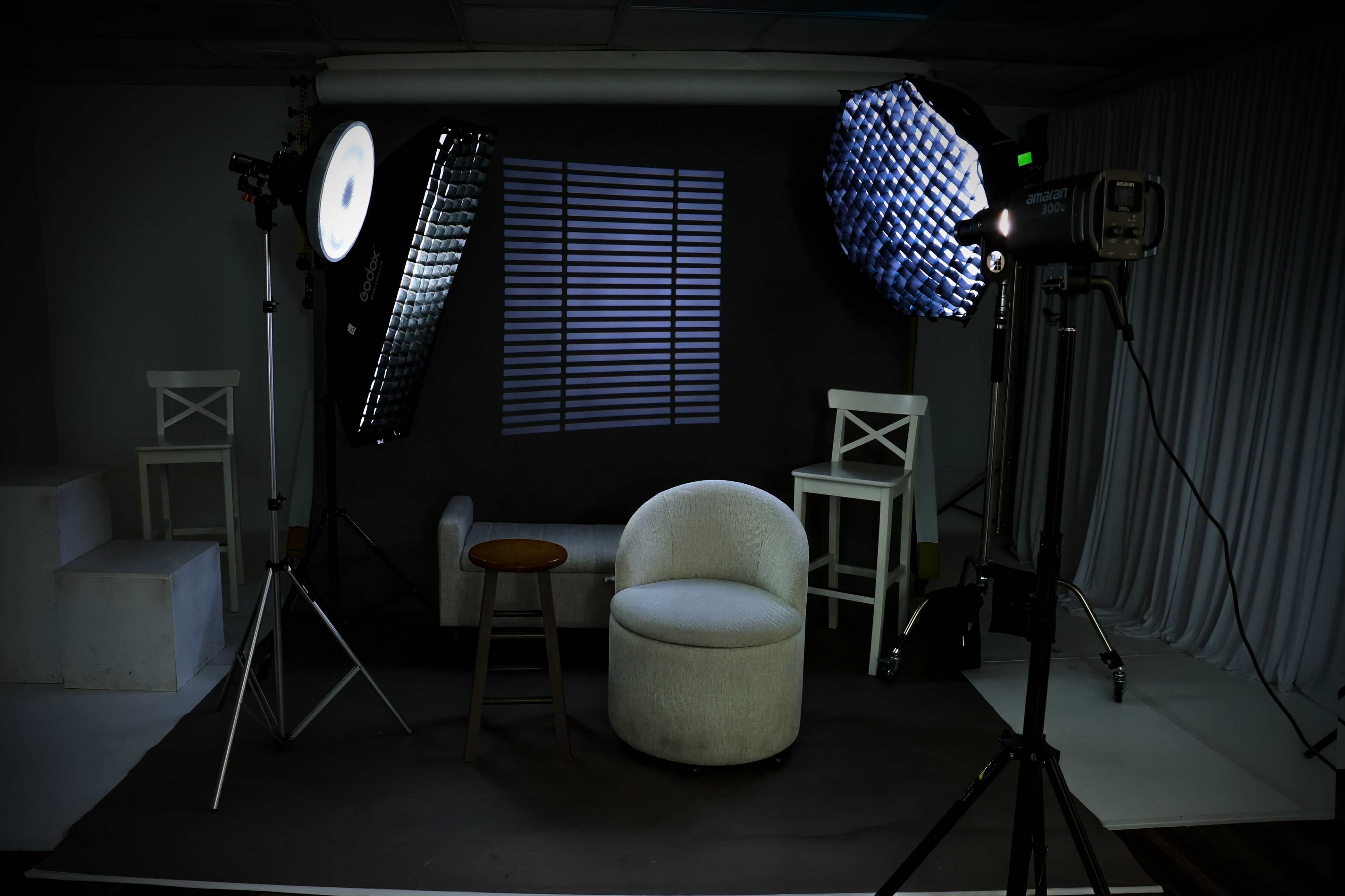 The image shows a photography studio setup with two light sources, a round chair, and a small stool against a backdrop of vertical blinds.