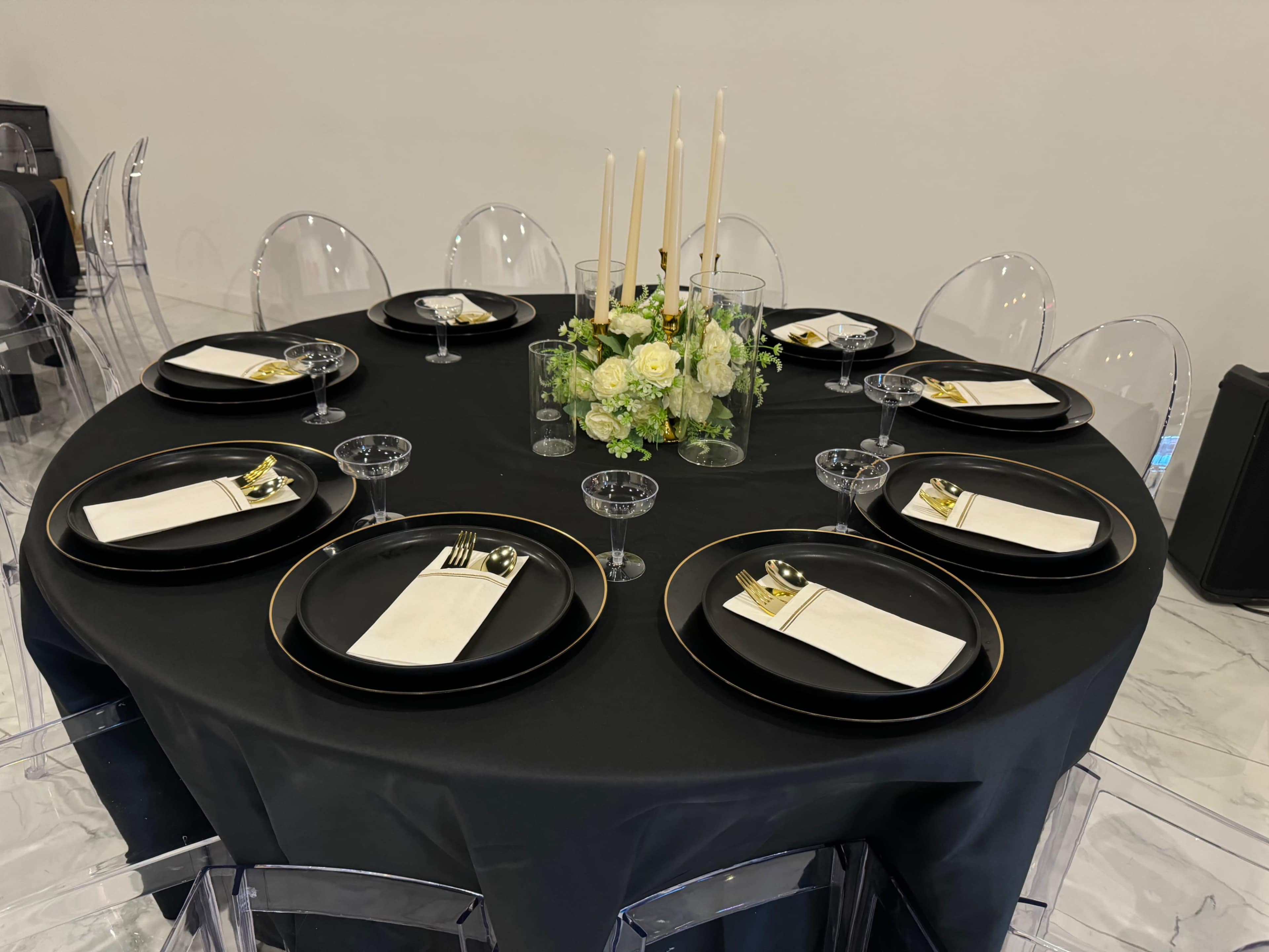 A round black table is set for dinner with clear chairs, elegant tableware, and a floral centerpiece featuring white roses and tall candles.