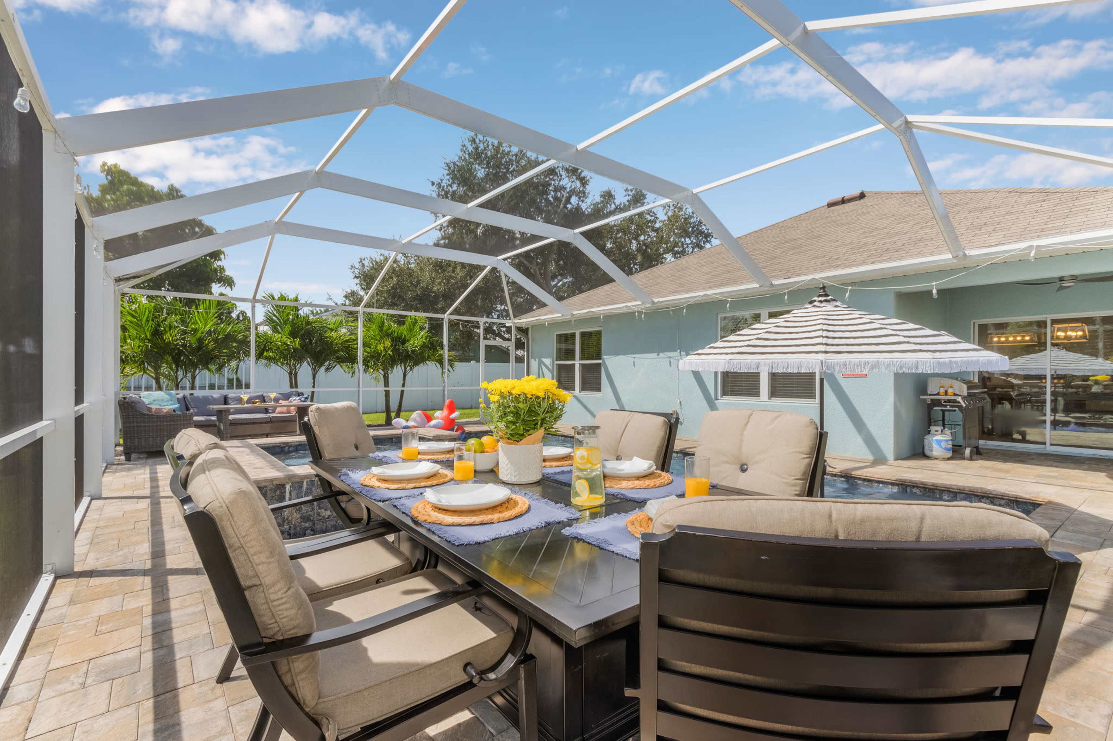 A screened patio features a dining table set with plates and drinks, surrounded by cushioned chairs and tropical plants, with a view of a pool.