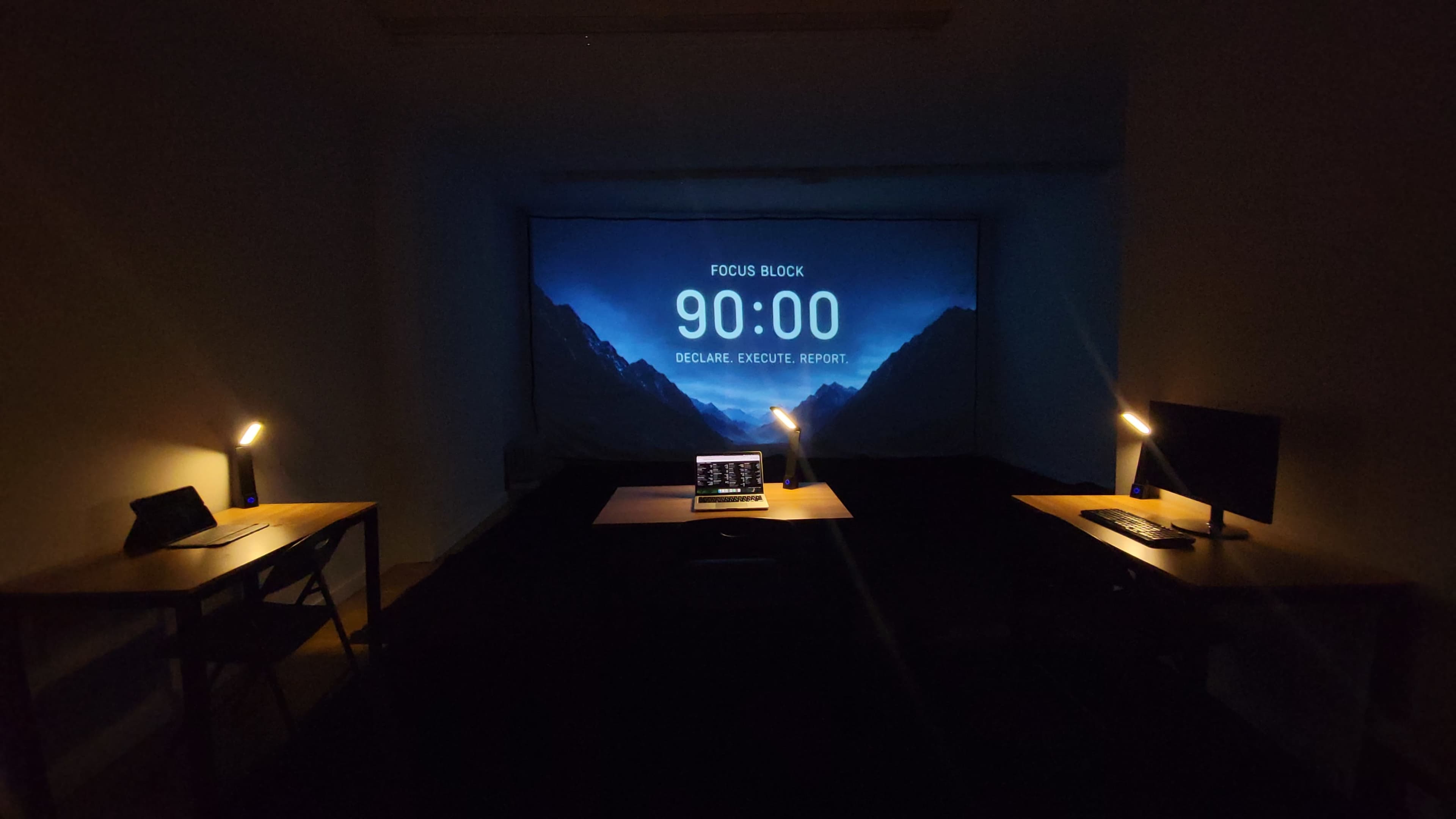 A dimly lit room features a large screen displaying a 90-minute timer, with two tables equipped with desk lamps and laptops on either side.
