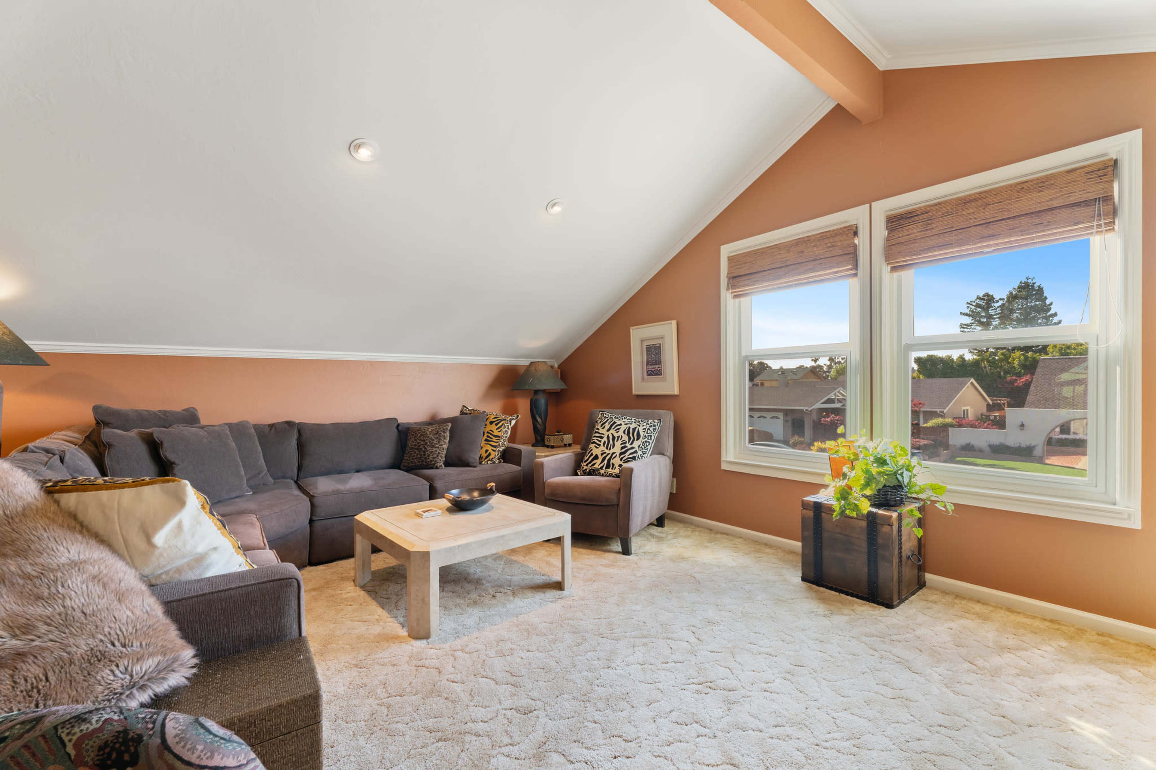 A well-lit attic living space featuring a gray sectional sofa, a matching armchair, a coffee table, and windows overlooking a residential area.