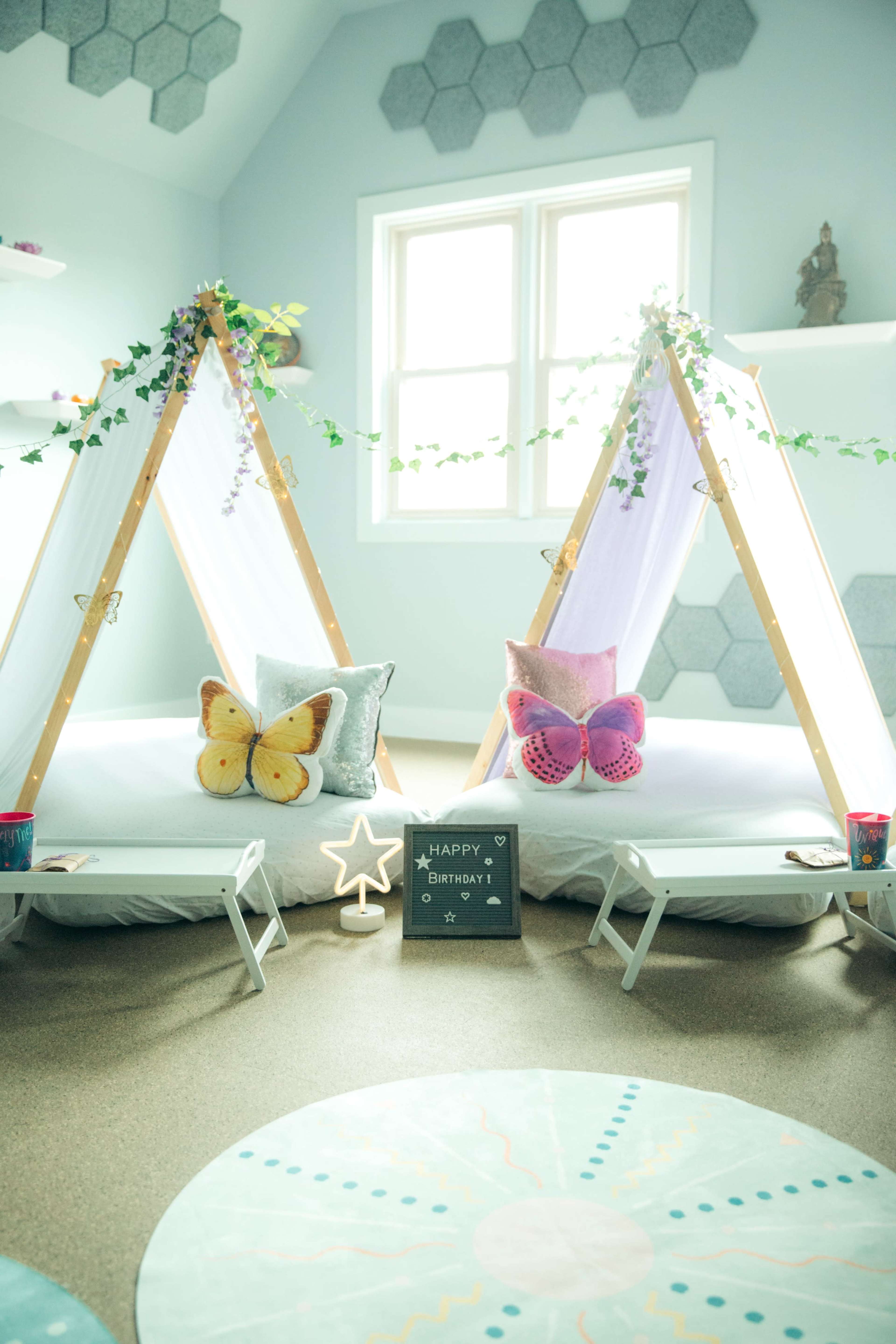 A decorated room with two children's play tents, adorned with pillows featuring butterfly designs, and a "Happy Birthday" sign positioned on the floor.