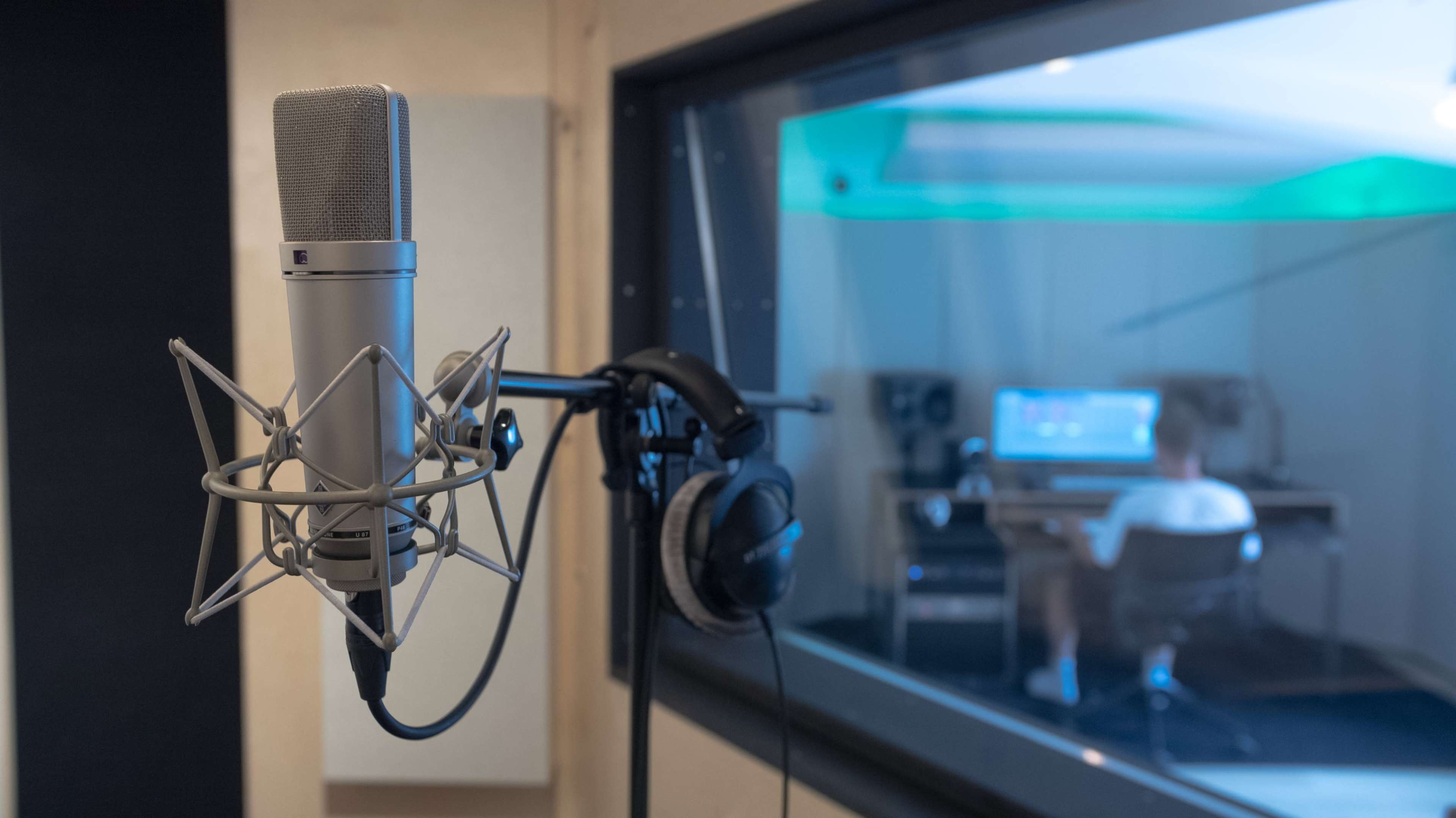 The image shows a microphone in the foreground with headphones attached, while a person works at a computer in a sound recording studio in the background.