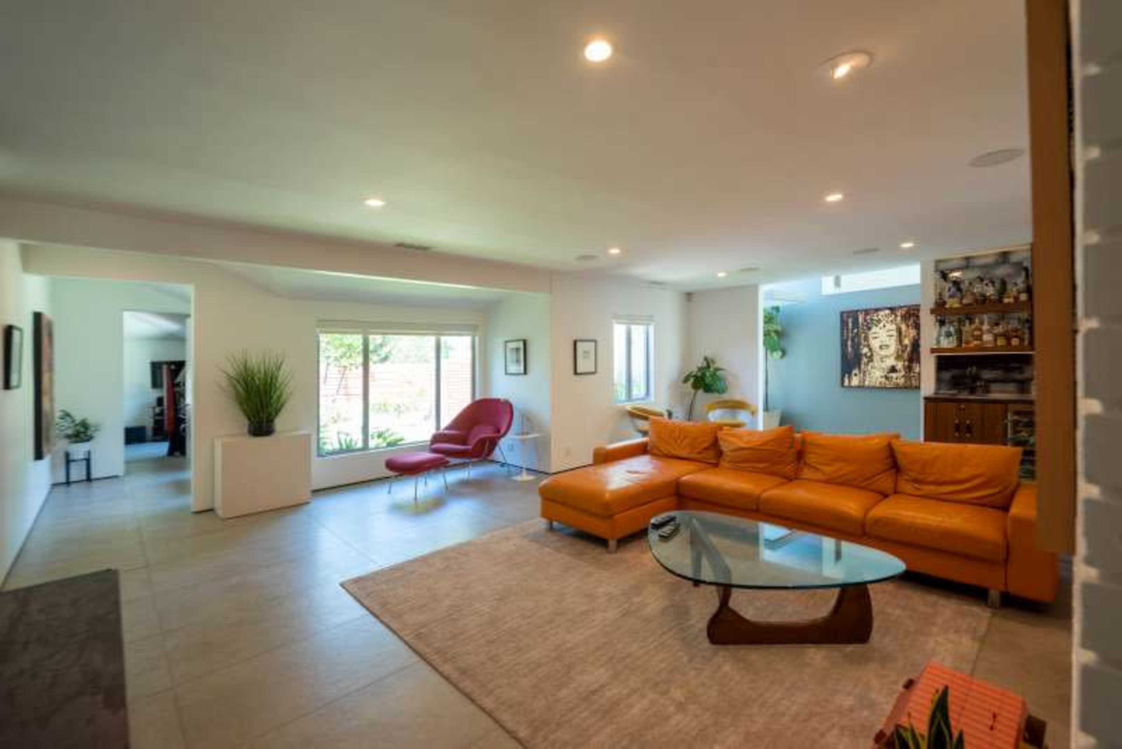 A modern living room with a large orange sectional sofa, a glass coffee table, and large windows allowing natural light to fill the space.