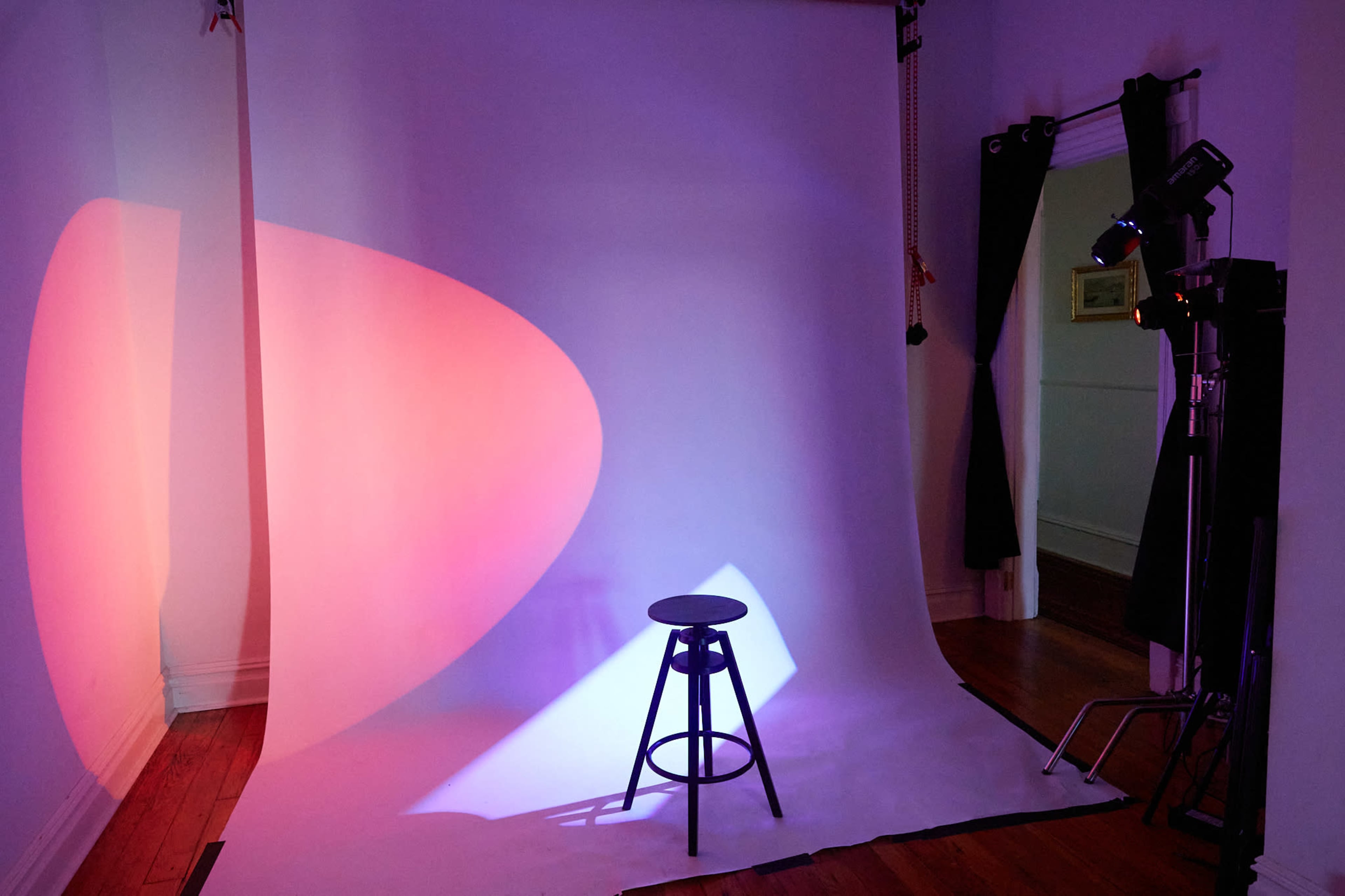 A dimly lit photography studio features a white backdrop with colorful lighting and a black stool positioned in front.