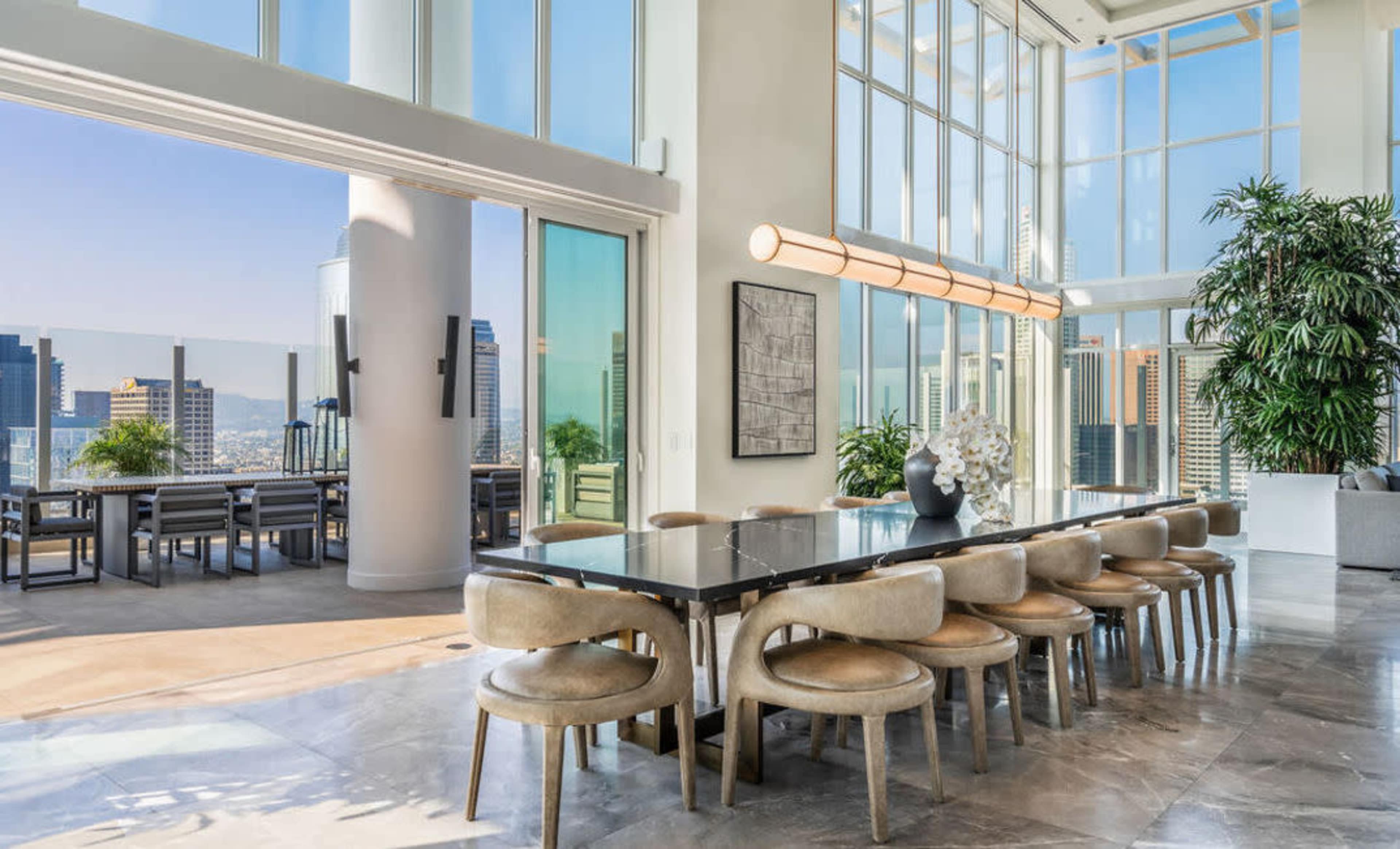 A spacious dining area with a long table and modern chairs overlooks a city skyline through large glass windows.